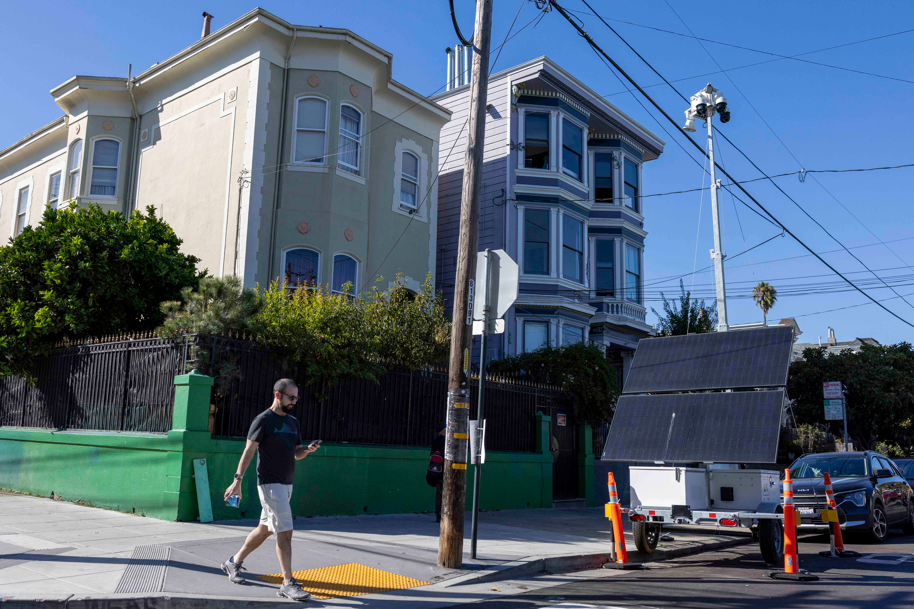 A man walks while looking at his phone on a sidewalk beside two multi-story houses; nearby, a pole with surveillance cameras and a portable solar panel setup is visible.