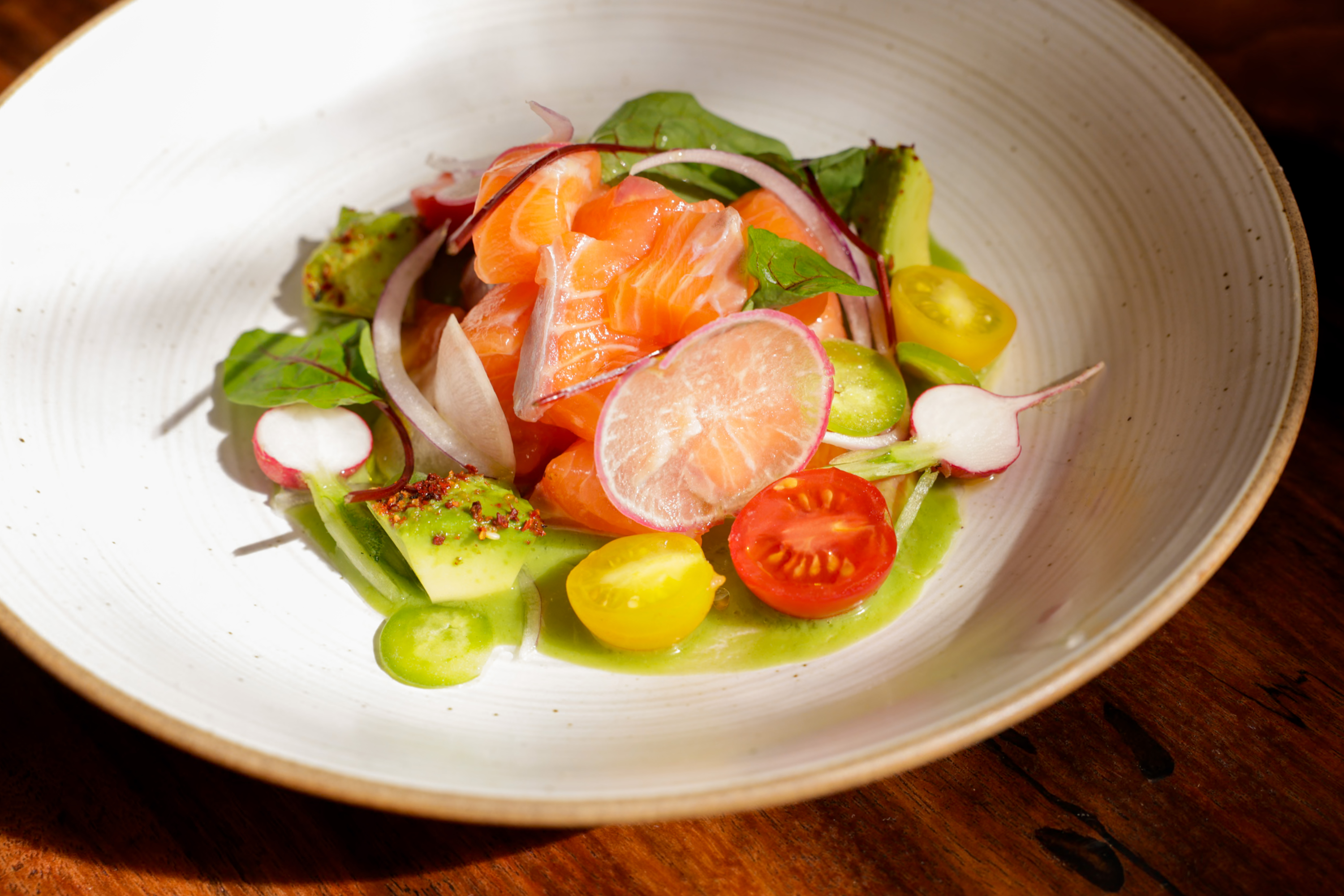 A white plate holds a vibrant salad with slices of salmon, radishes, cherry tomatoes, avocado, onion, and greens, all lightly drizzled with a green sauce.