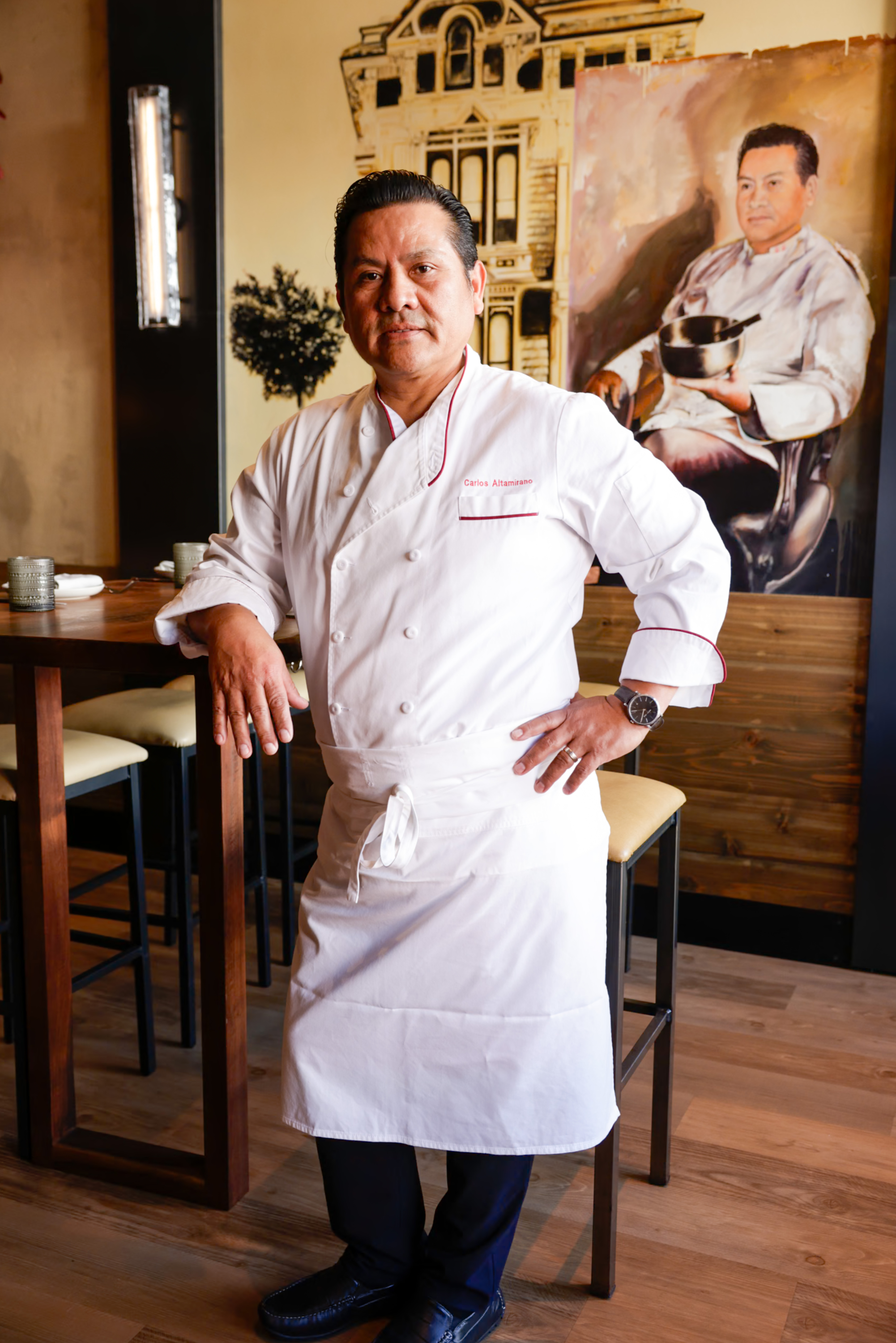 A chef in a white coat stands confidently beside a wooden table. Behind him, there's a portrait featuring the chef holding a pot, with a vintage building in the background.