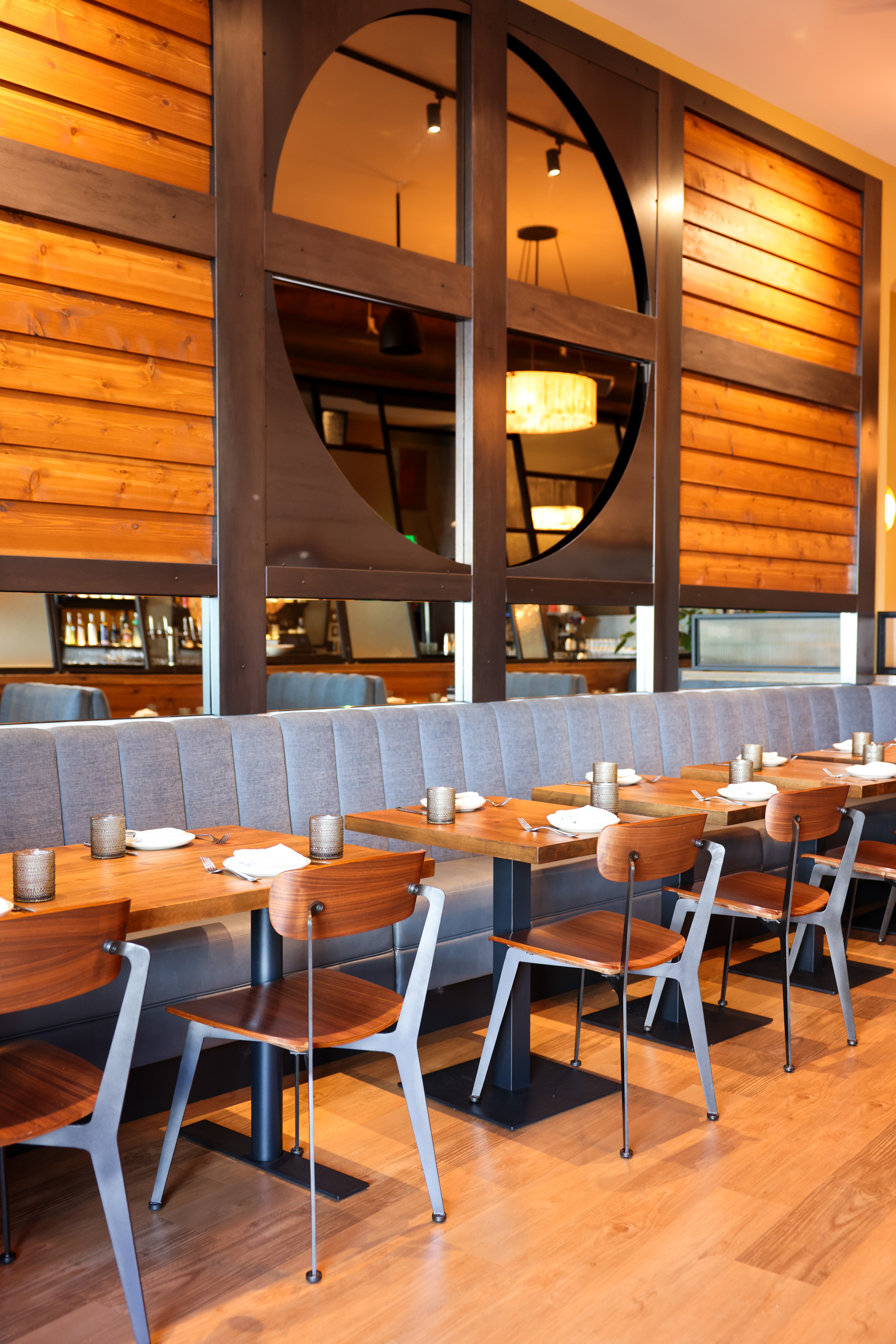 The image shows a modern restaurant with wooden chairs, matching tables set with napkins, and a blue cushioned bench against a wood and metal partition.