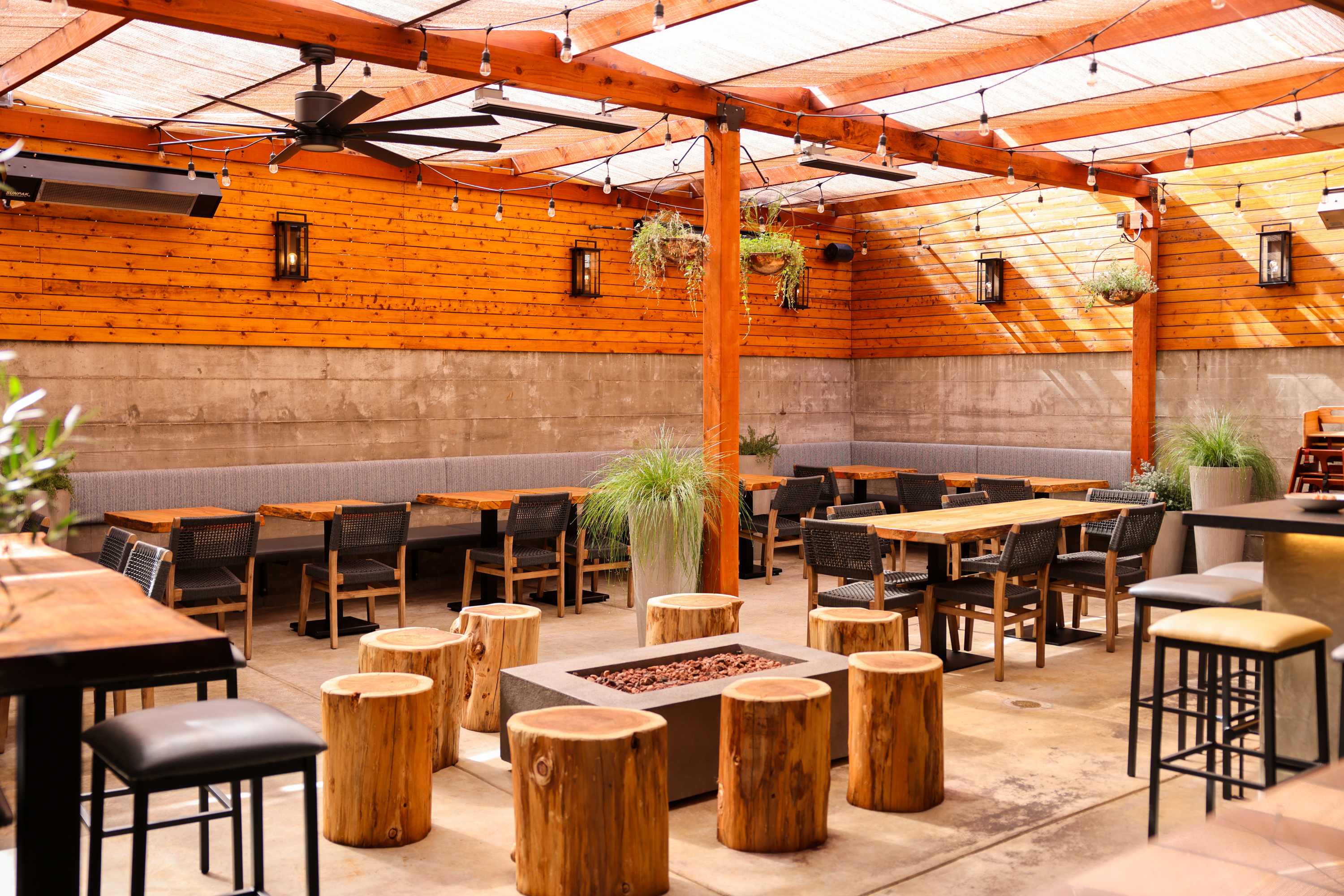 The image shows a cozy patio with wooden furniture, a fire pit surrounded by log stools, tables with chairs, and hanging plants under a pergola with string lights.