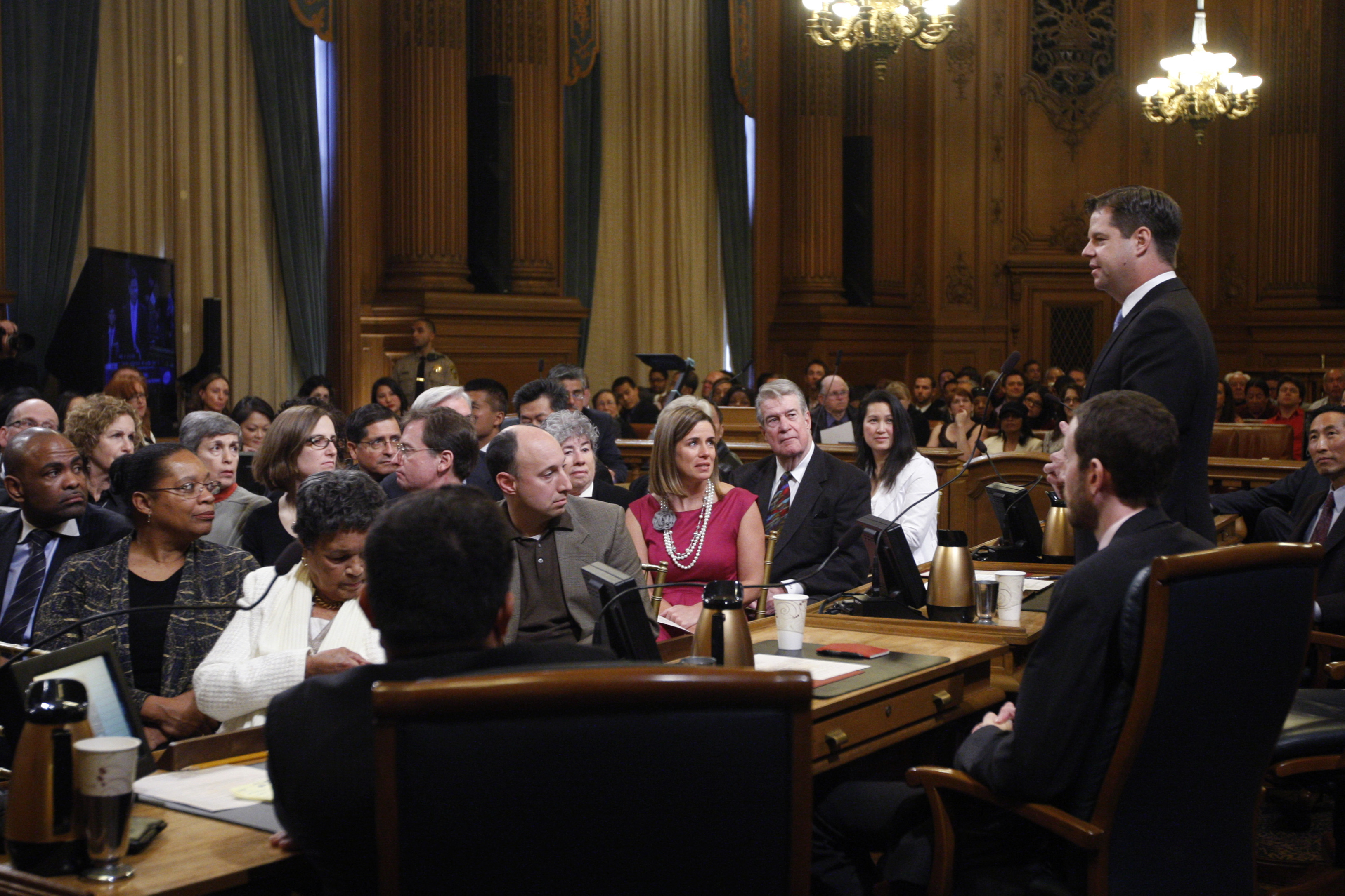 A caucasian man on the right in a suit takes an oath of office with his hand raised before a crowd in an ornate wood room.