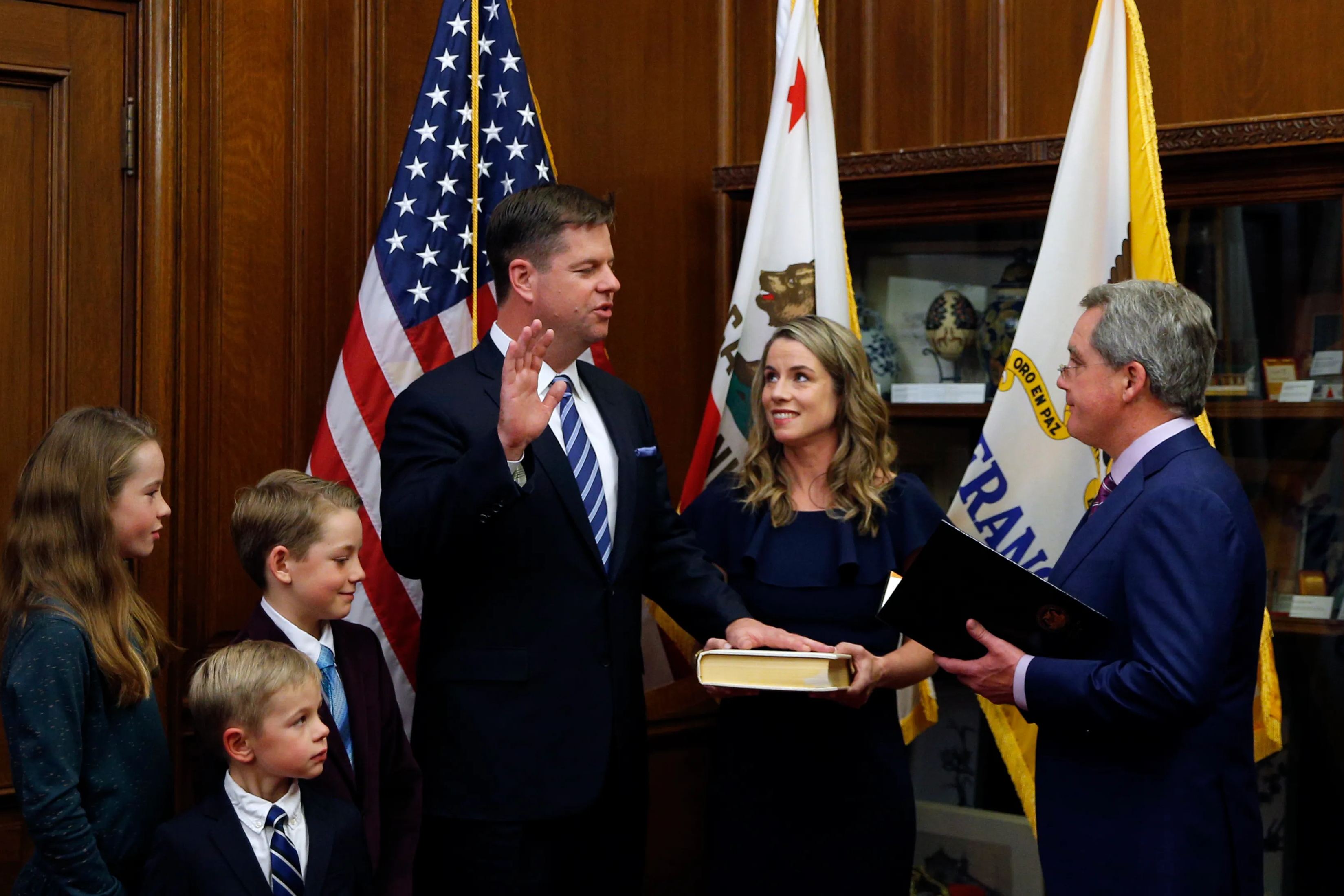 Mark Farrell, a white man in a suit, stands on the left side of the frame surrounded by his two kids and wife, as he takes an oath of office with a hand raised.