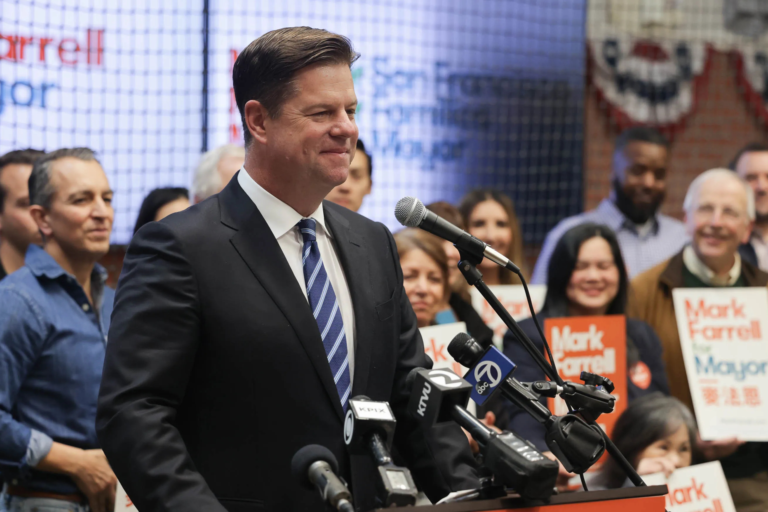 A man in a suit stands at a podium with multiple microphones, smiling, surrounded by a crowd holding signs. The background includes a blurred campaign sign.