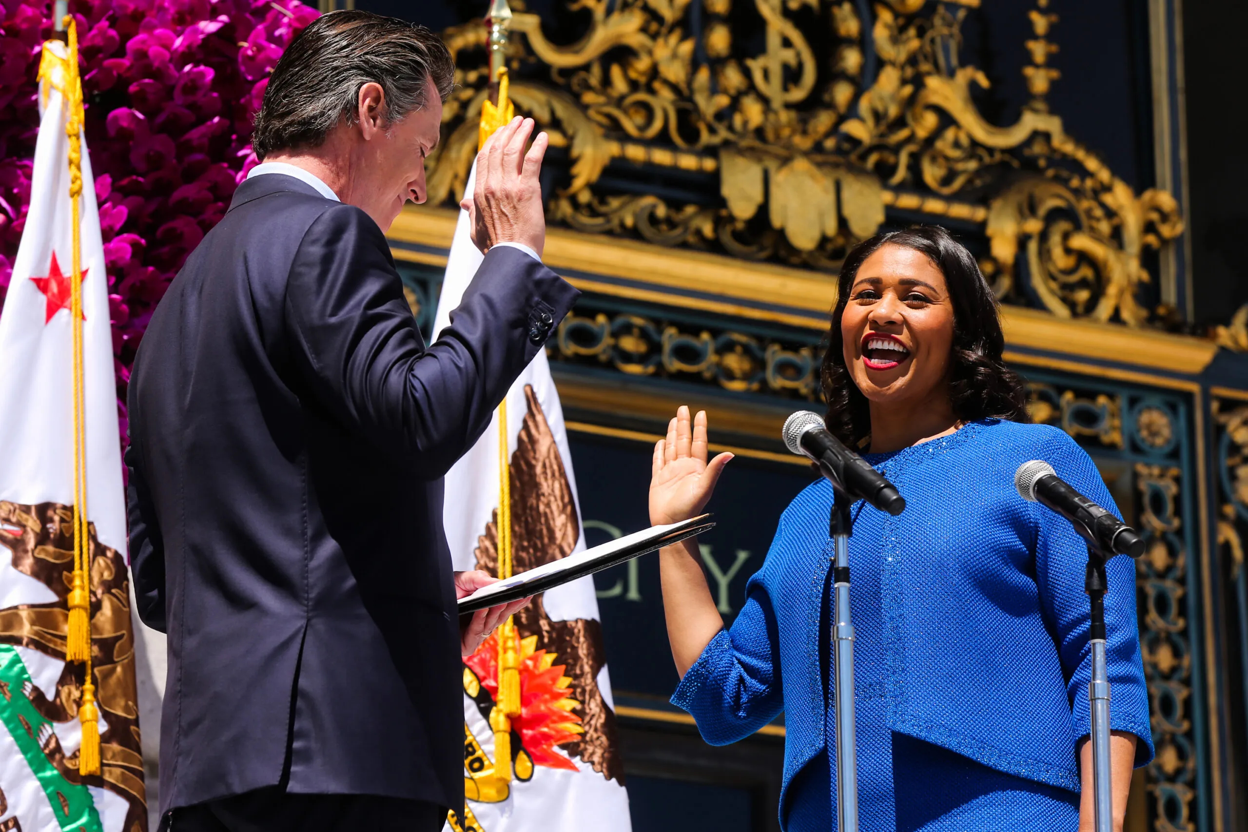 London Breed, a Black woman in a blue suit holds her hand up to take the oath of office in front of city hall, facing Gavin Newsom, a white man, whose back is turned.