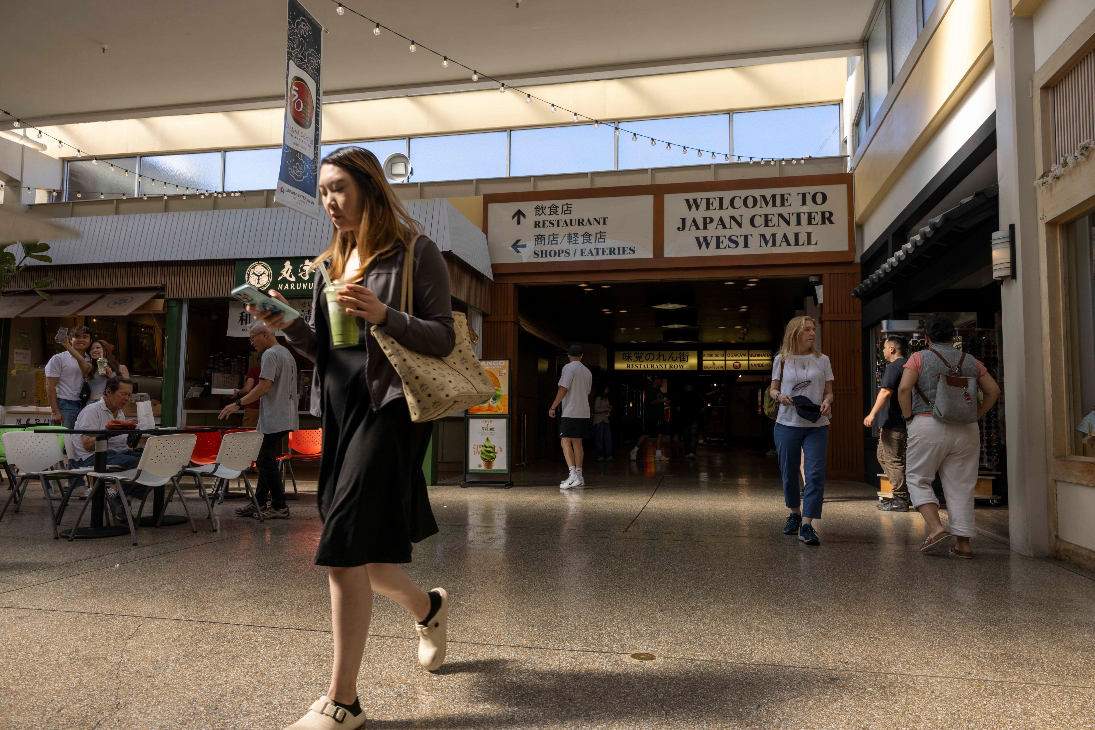 A woman walks through a mall, holding a drink and checking her phone. The sign above reads "Welcome to Japan Center West Mall," with people and shops in the background.