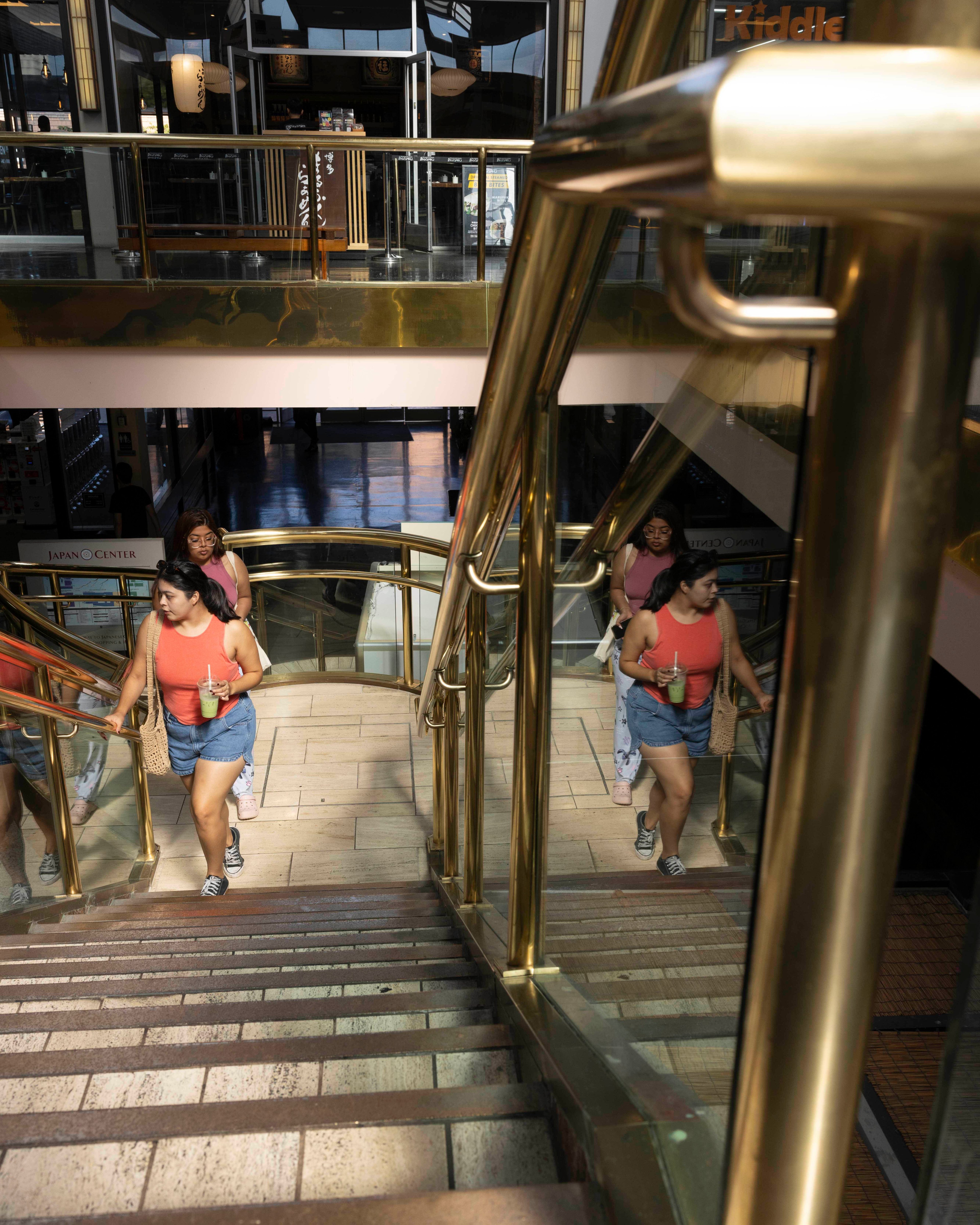 Two people walk up a staircase with gold railings in a mall. One wears a red top and denim shorts, holding a drink, while their reflections appear on the glass sides.