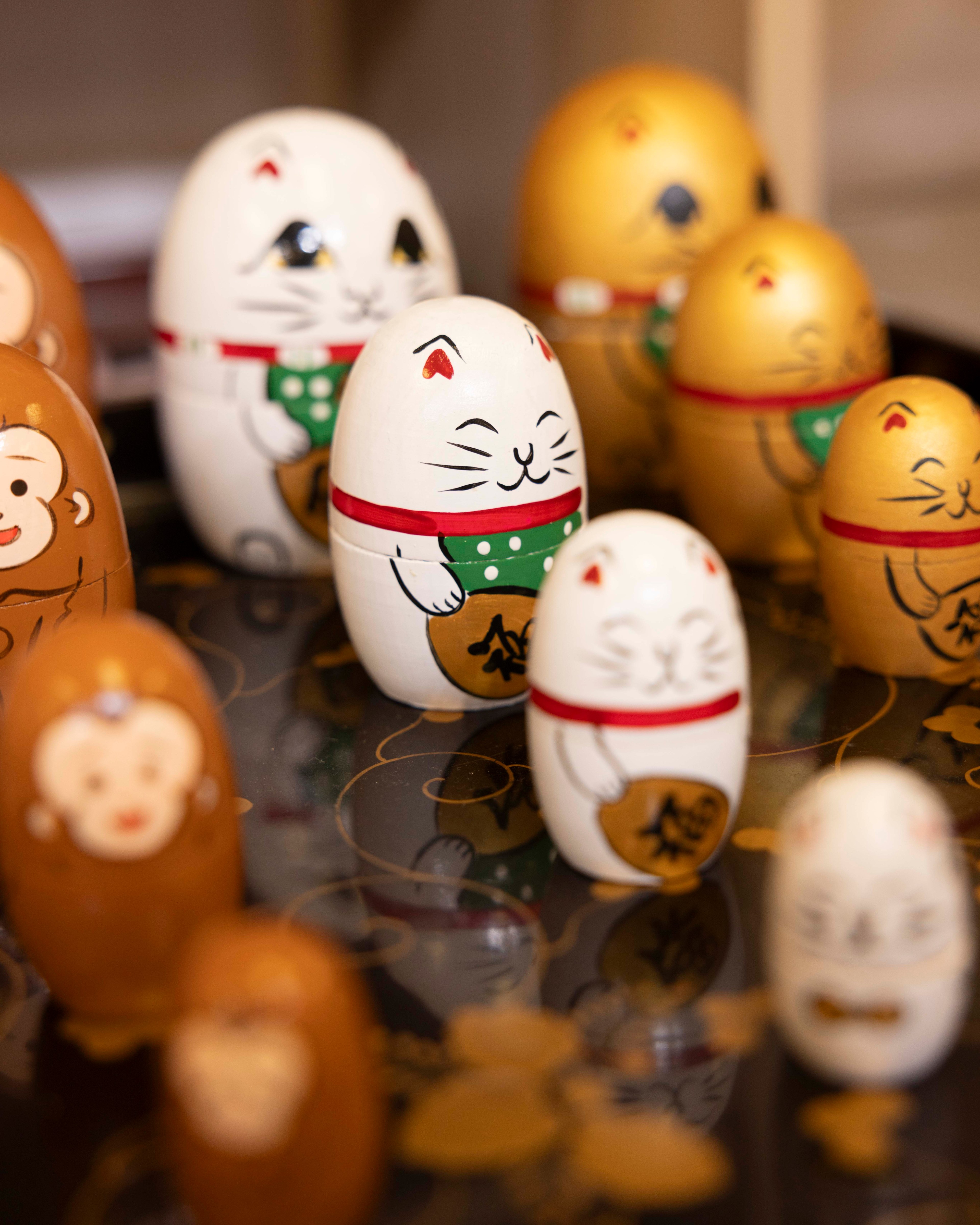 The image depicts decorative wooden dolls shaped like cats and monkeys. The cats are painted white with red collars and hold a gold coin. The background has a dark floral pattern.