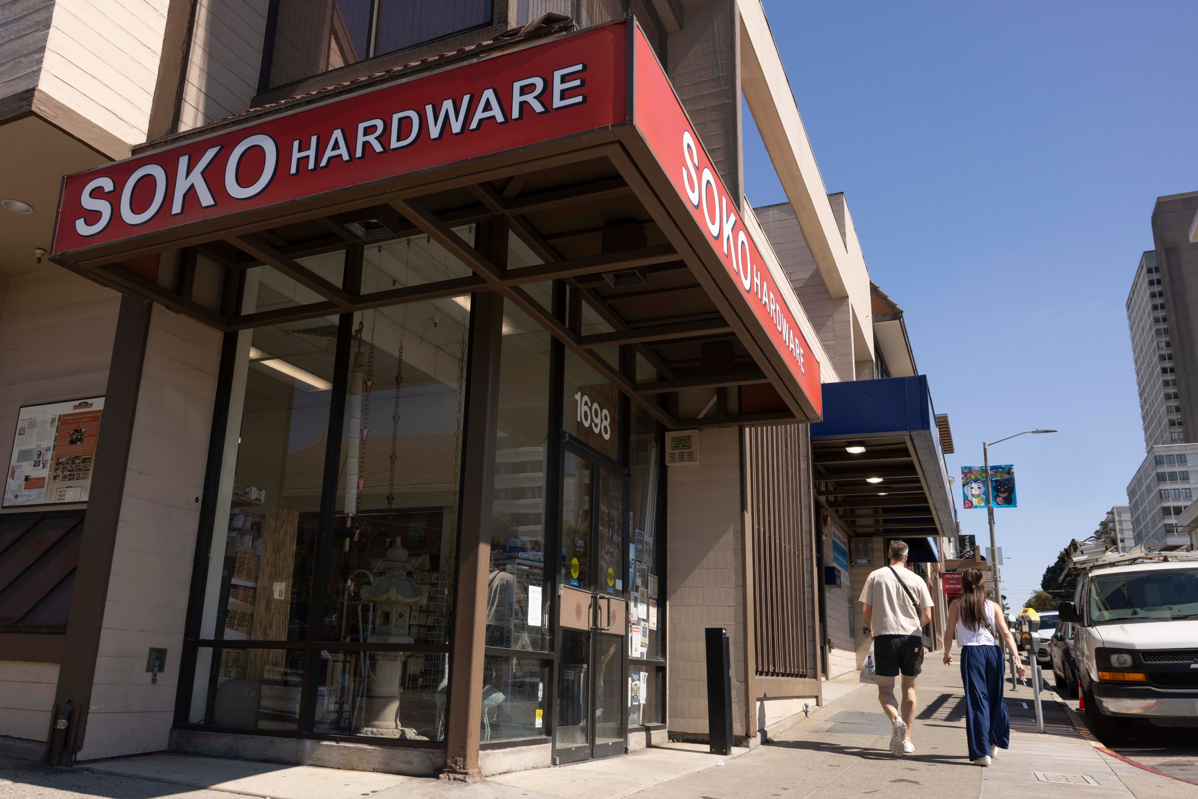 A street scene shows a couple walking past Soko Hardware, a store with a red sign. The street is lined with buildings, a utility van, and a clear blue sky above.