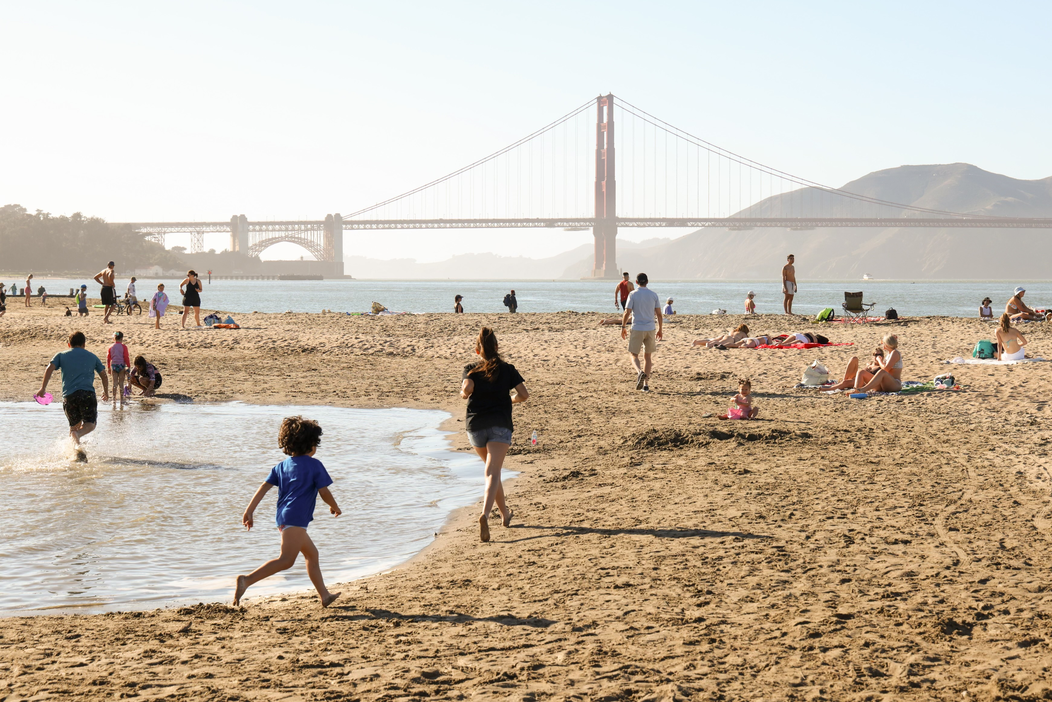 People relax on a sunny beach with the Golden Gate Bridge in the background. Children play near a shallow water area. Some are sunbathing on the sand.