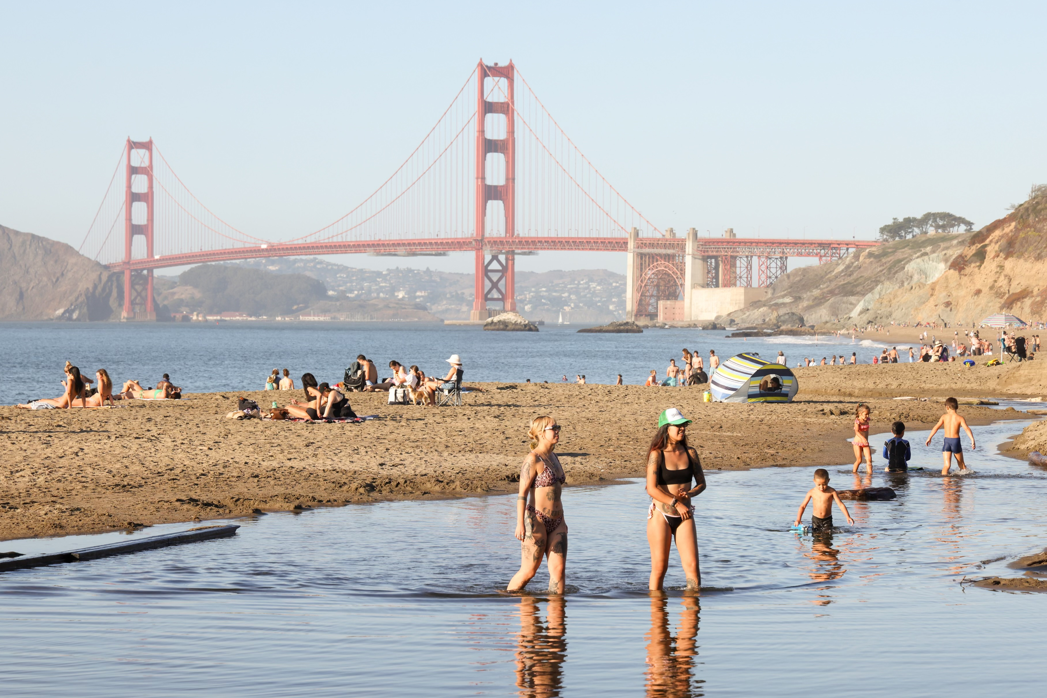 People relax on a beach with the Golden Gate Bridge in the background. Some are sunbathing, while others, including children, wade in shallow water.