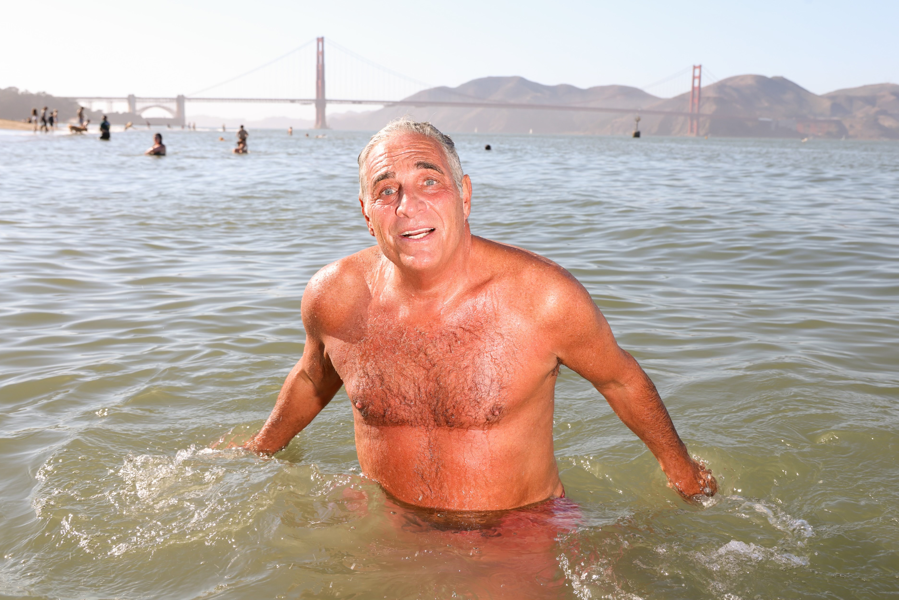 A man stands in the water with a joyful expression. In the background, there's a view of the Golden Gate Bridge and distant hills, with people swimming nearby.