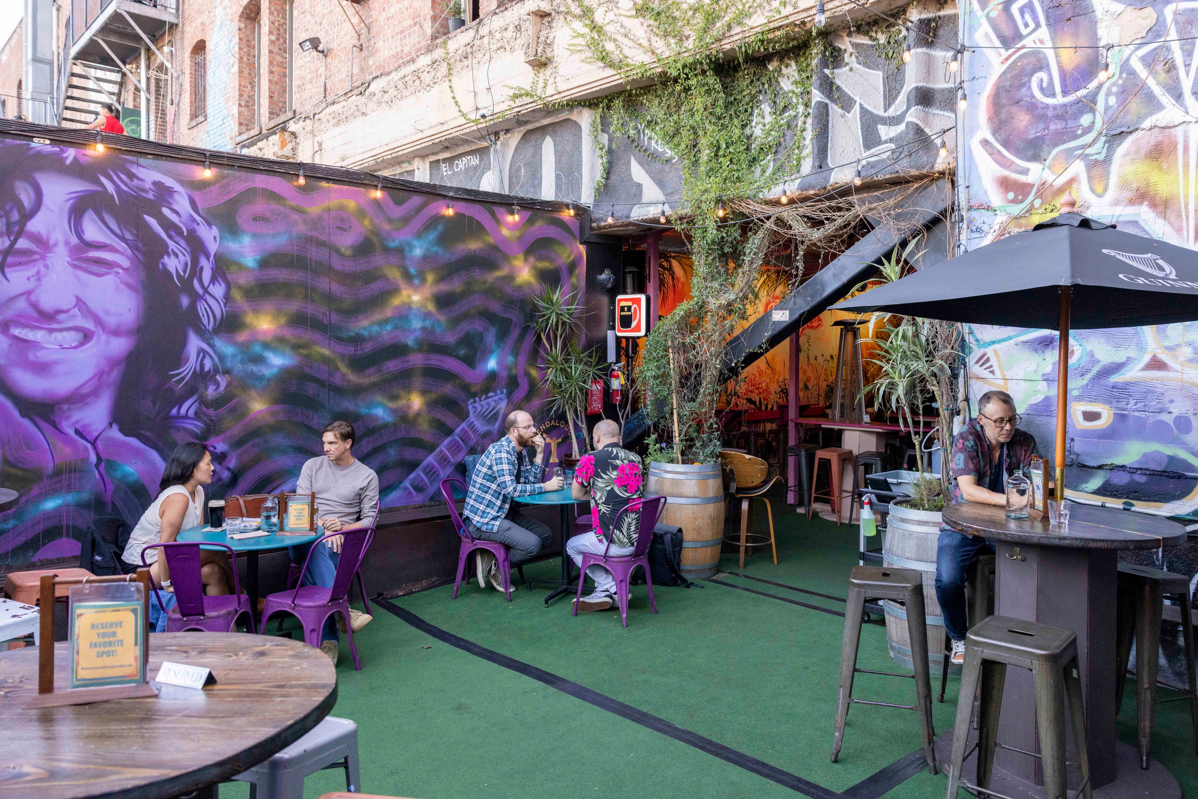 People sit at outdoor tables against a mural-covered wall, under string lights. The space features plants, a staircase, and a relaxed, artsy atmosphere.