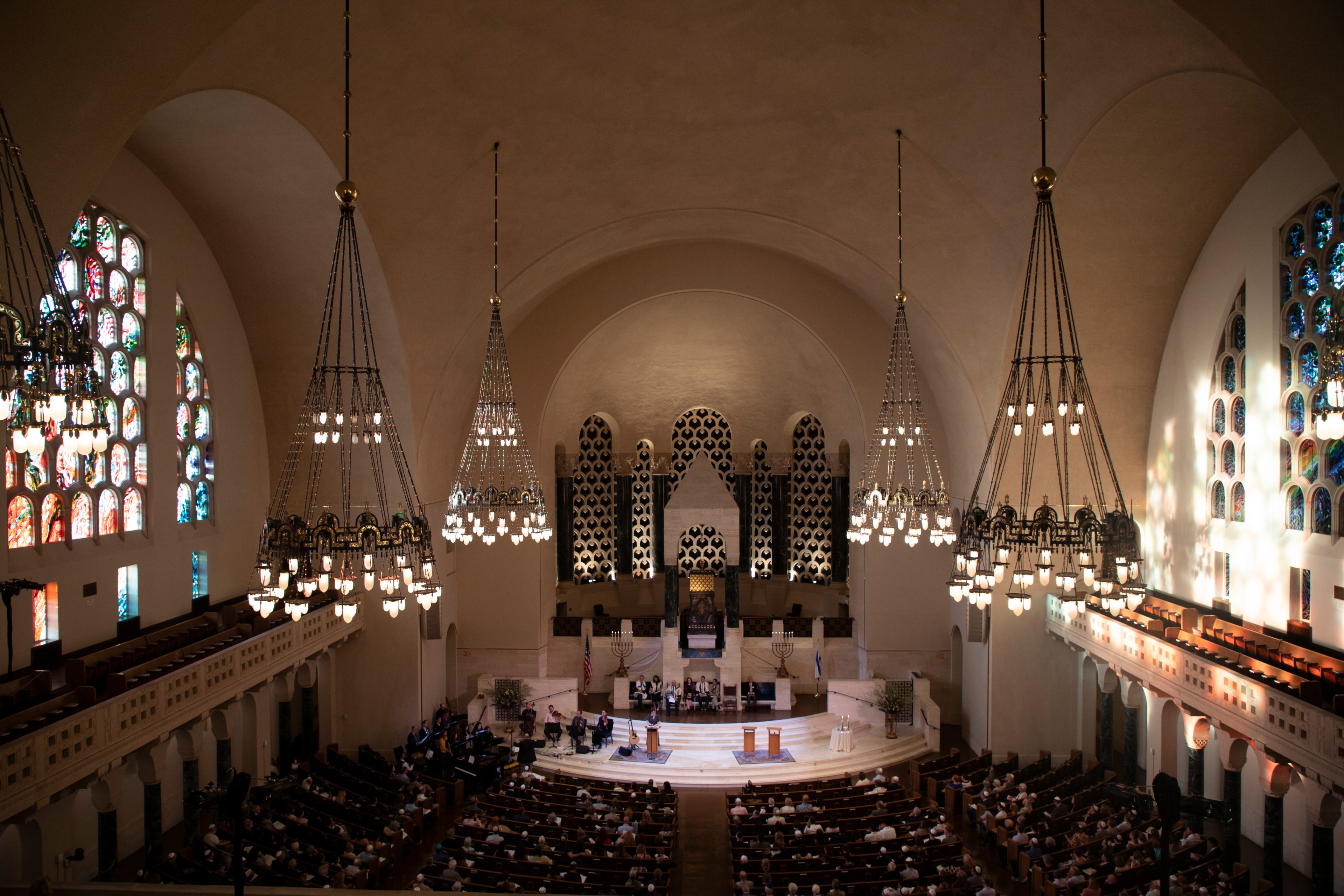 A large synagogue with stained glass windows, grand chandeliers, and a stage. People are seated in pews, attending a service at the front where a choir is performing.