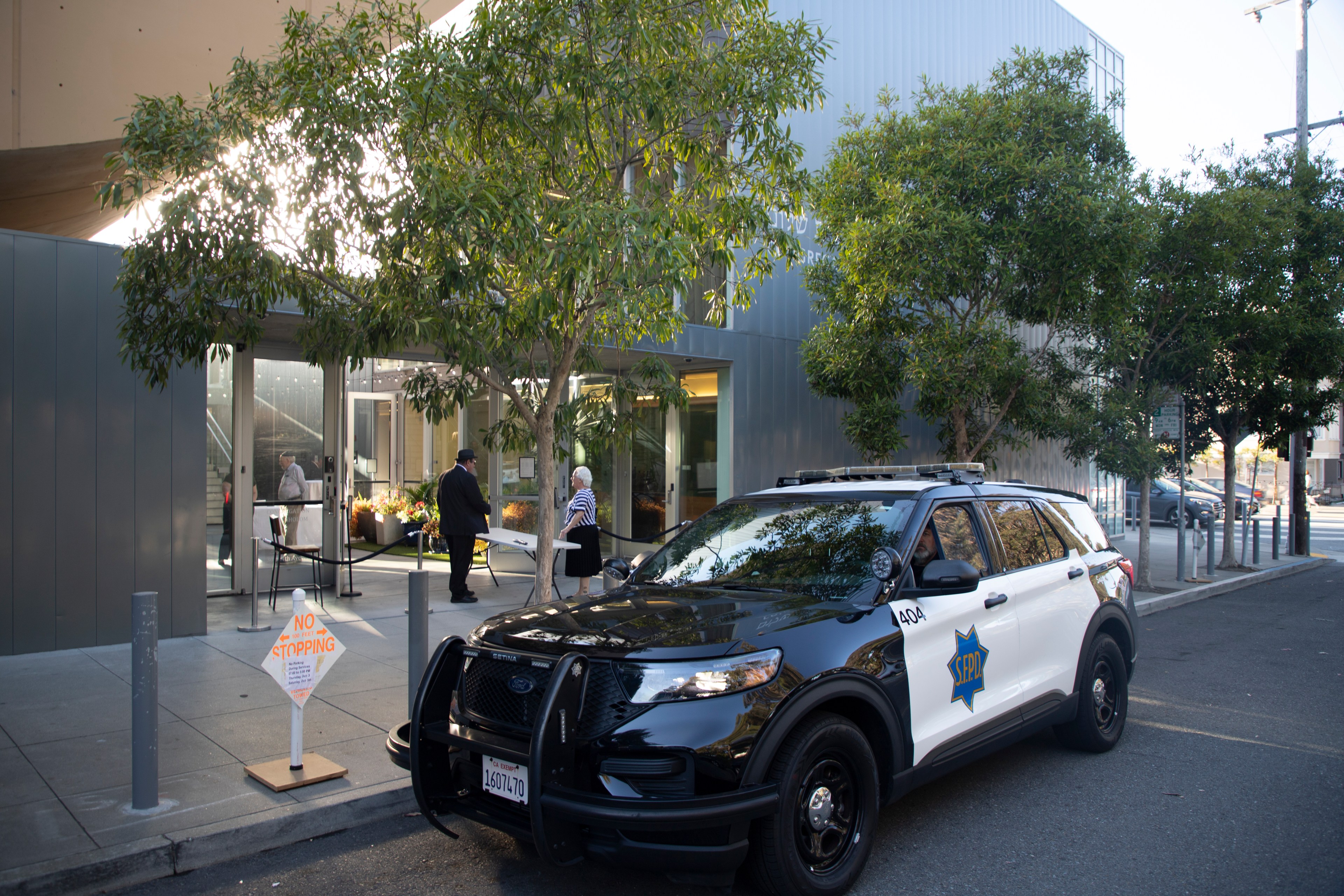 A black and white police SUV is parked on the street near a modern building entrance, with two people conversing under a tree. A &quot;No Stopping&quot; sign is visible.