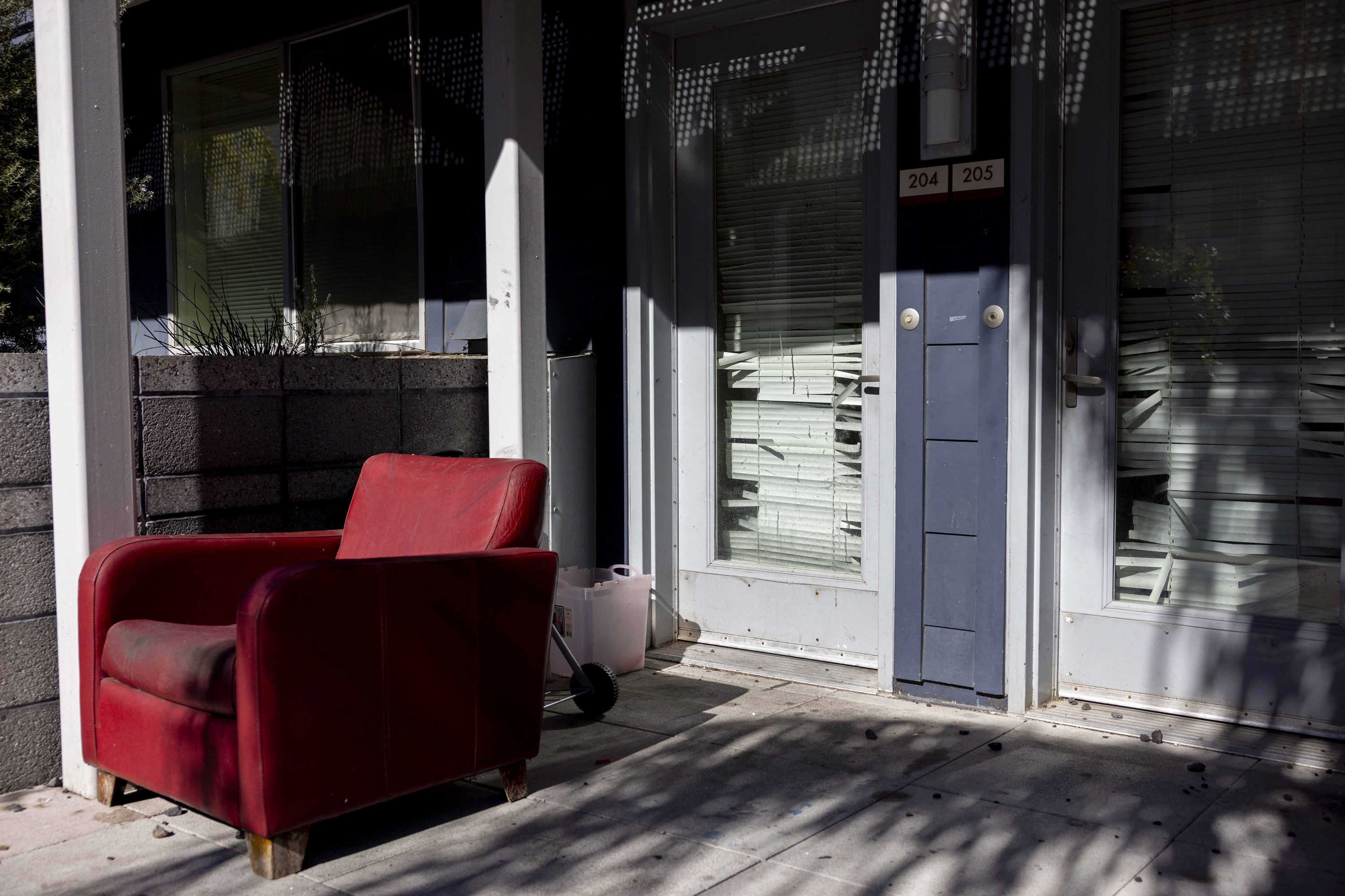 A red armchair sits outside near light blue doors with broken blinds. Numbers 204 and 205 are on the doors, and shadows stretch across the ground.