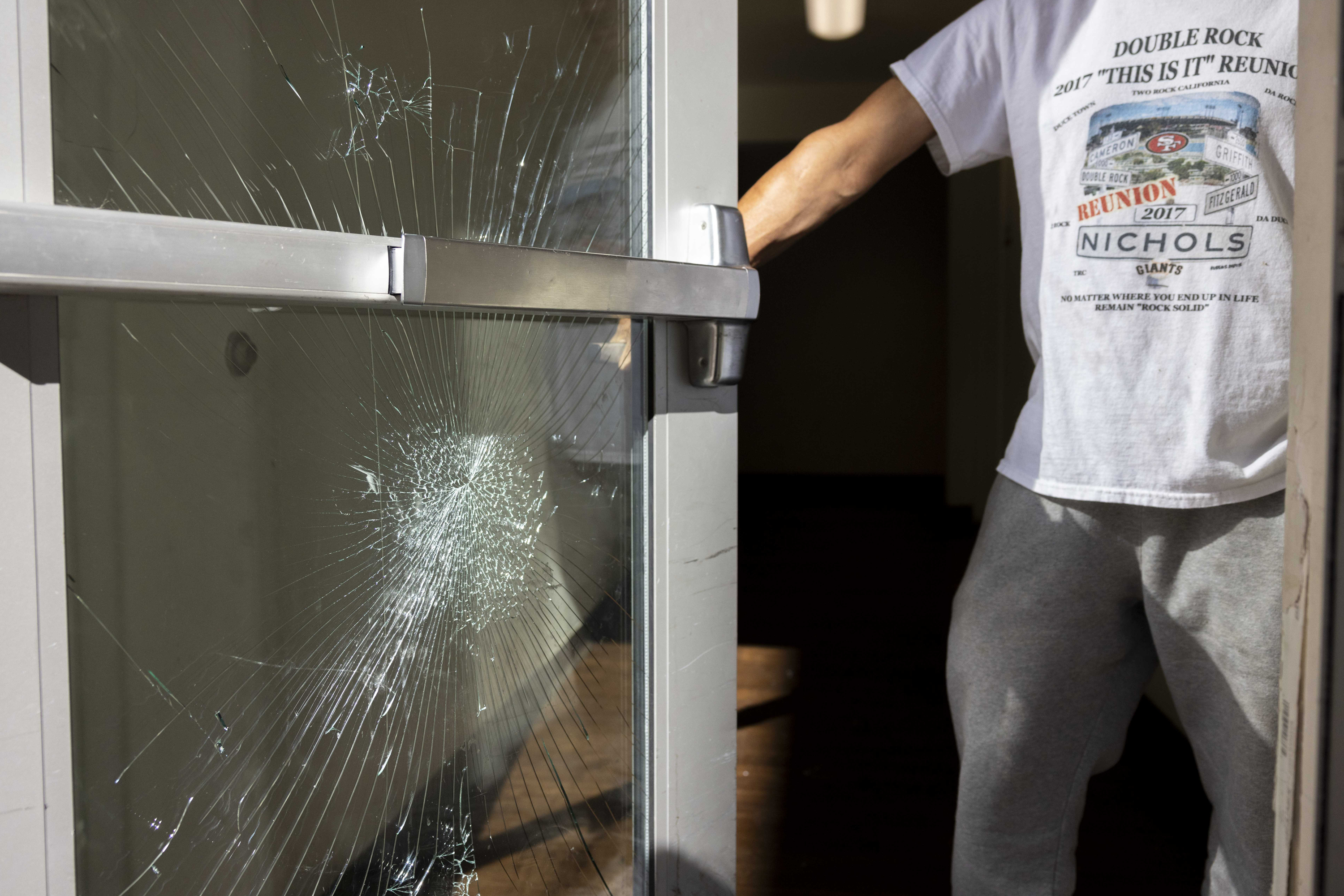 A person is pushing open a glass door with a large crack in it. They wear a white T-shirt and gray pants, and the door has a metal push bar.