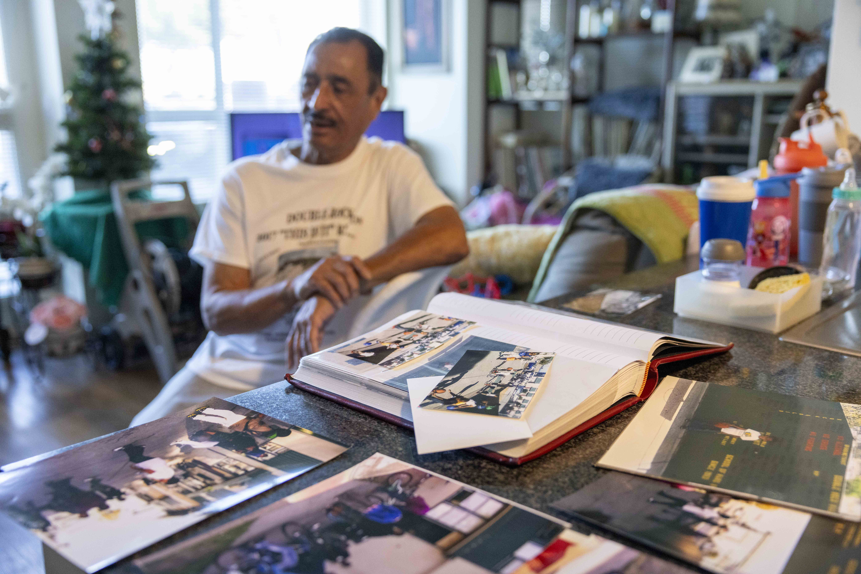 A man in a T-shirt sits near a table covered with scattered photos and an open photo album, in a cozy room with a small Christmas tree in the background.