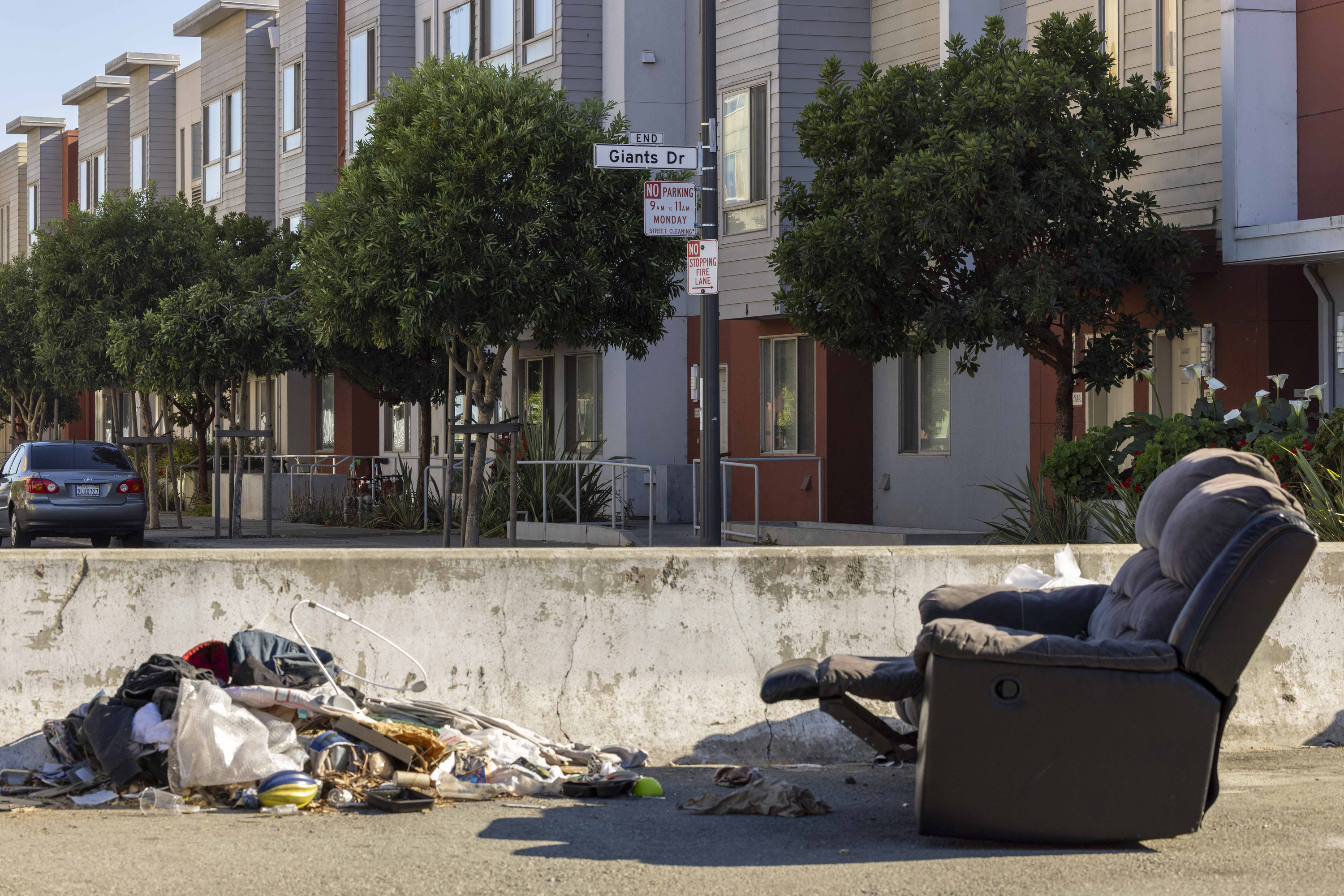 A pile of trash and a discarded recliner chair are set on the street in front of modern townhouses, with trees lining the sidewalk.