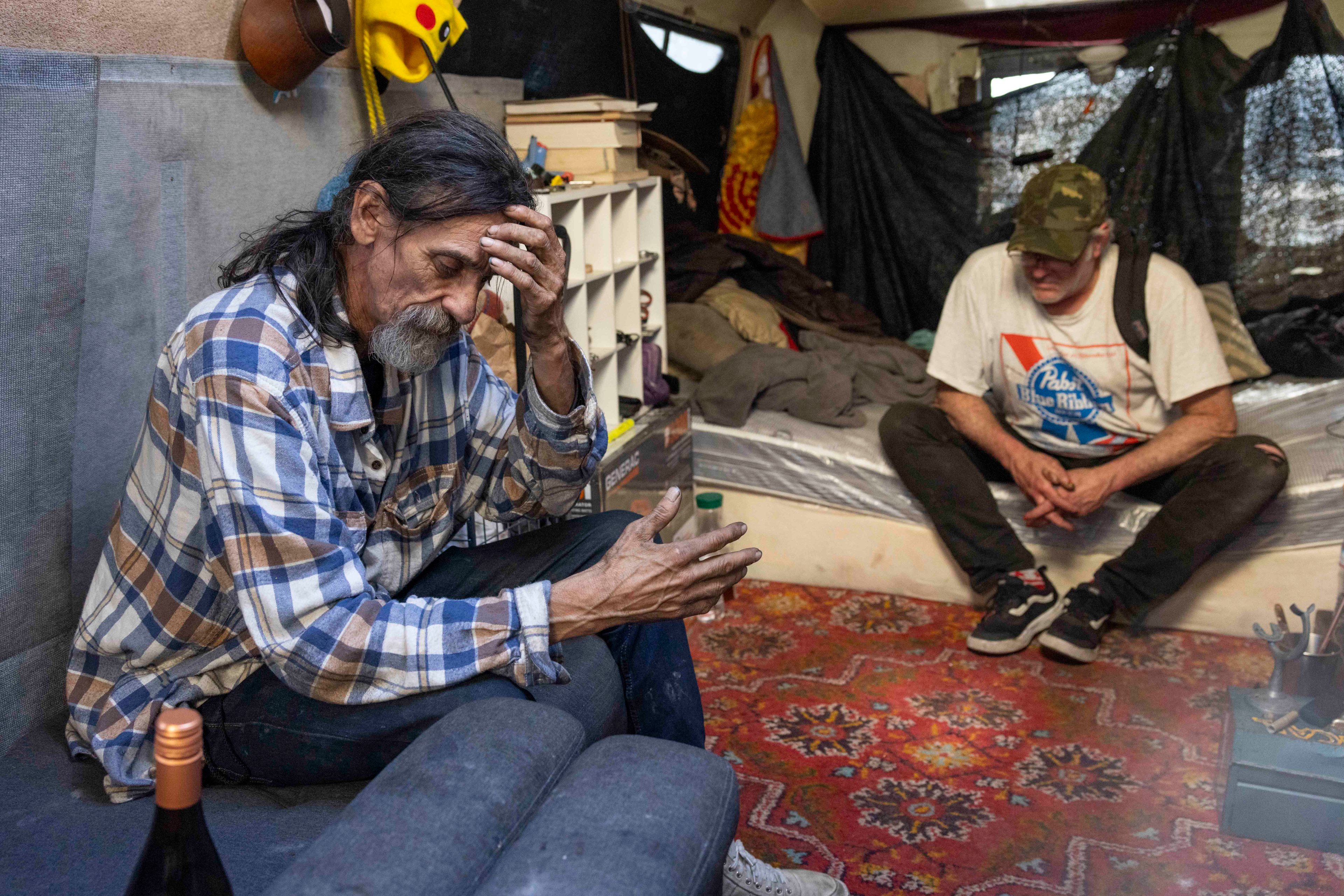 Two men sit in a cluttered, dimly lit room; one is on a sofa holding his head, the other on a bed wearing a cap. The room has a colorful carpet and scattered items.