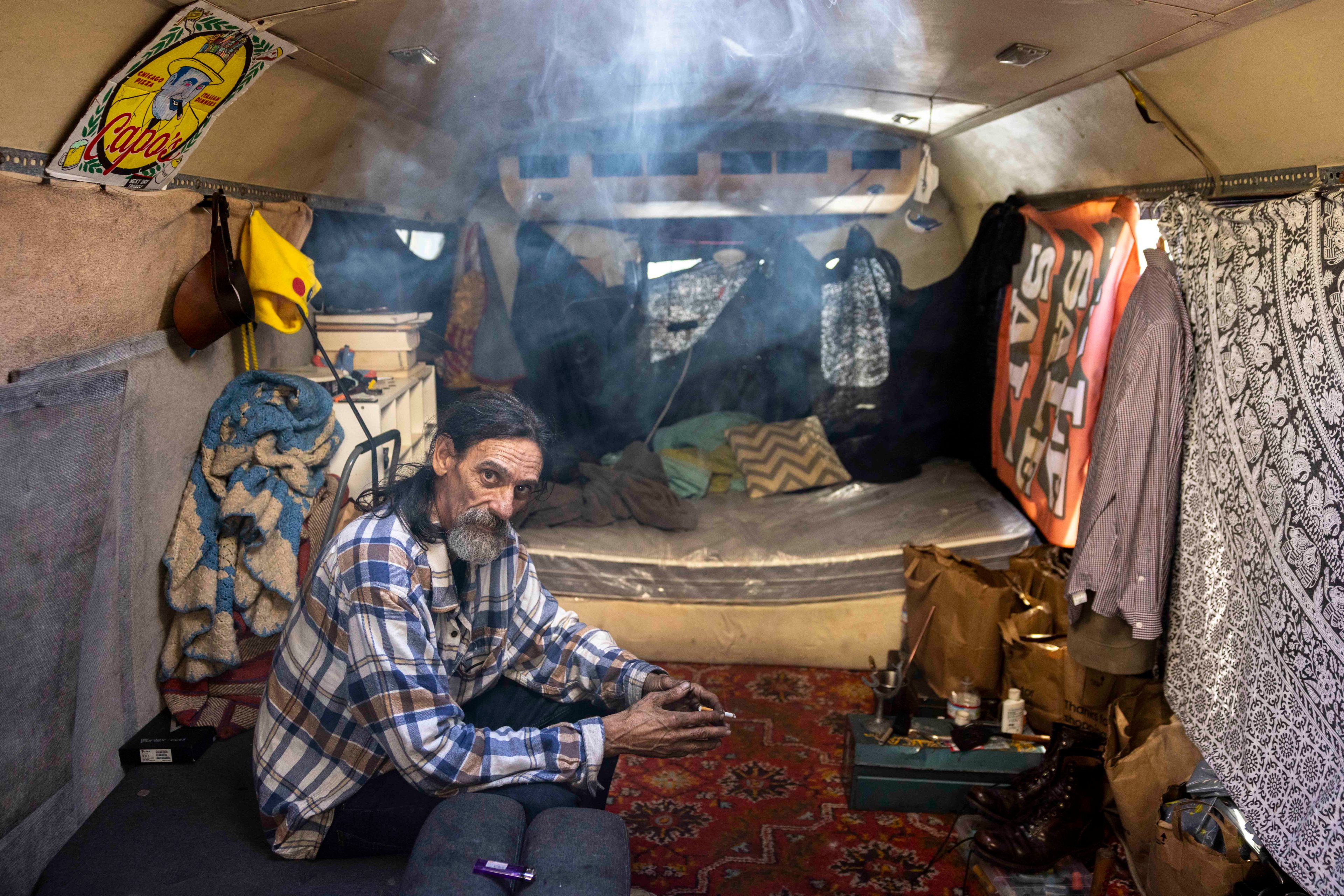 A man with a plaid shirt sits inside a cluttered camper with hanging clothes and blankets. Soft light filters through, creating a smoky, cozy atmosphere.