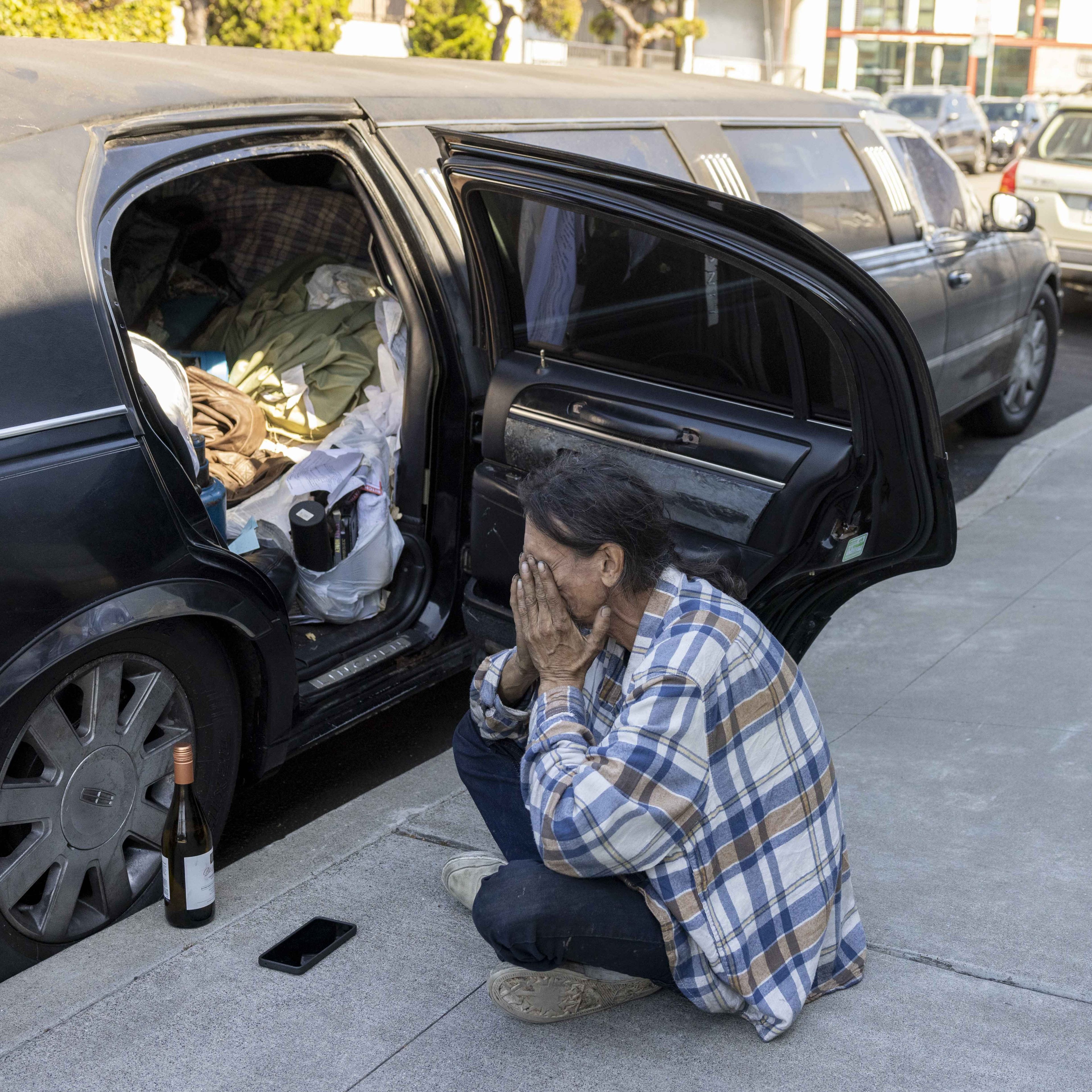 A person in a plaid shirt crouches by an open car door filled with belongings. A bottle and a phone are on the ground in front of them.