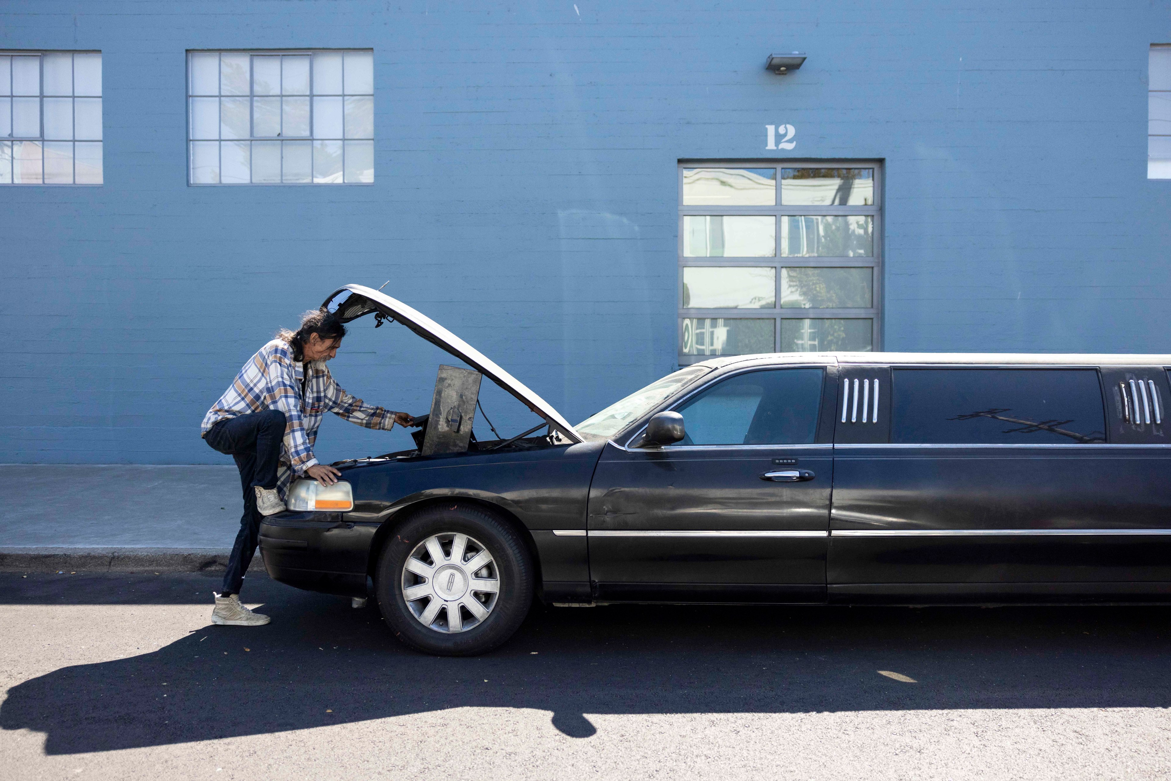 A person in a plaid shirt works under the open hood of a black limousine parked on a street, with a blue industrial building numbered &quot;12&quot; in the background.