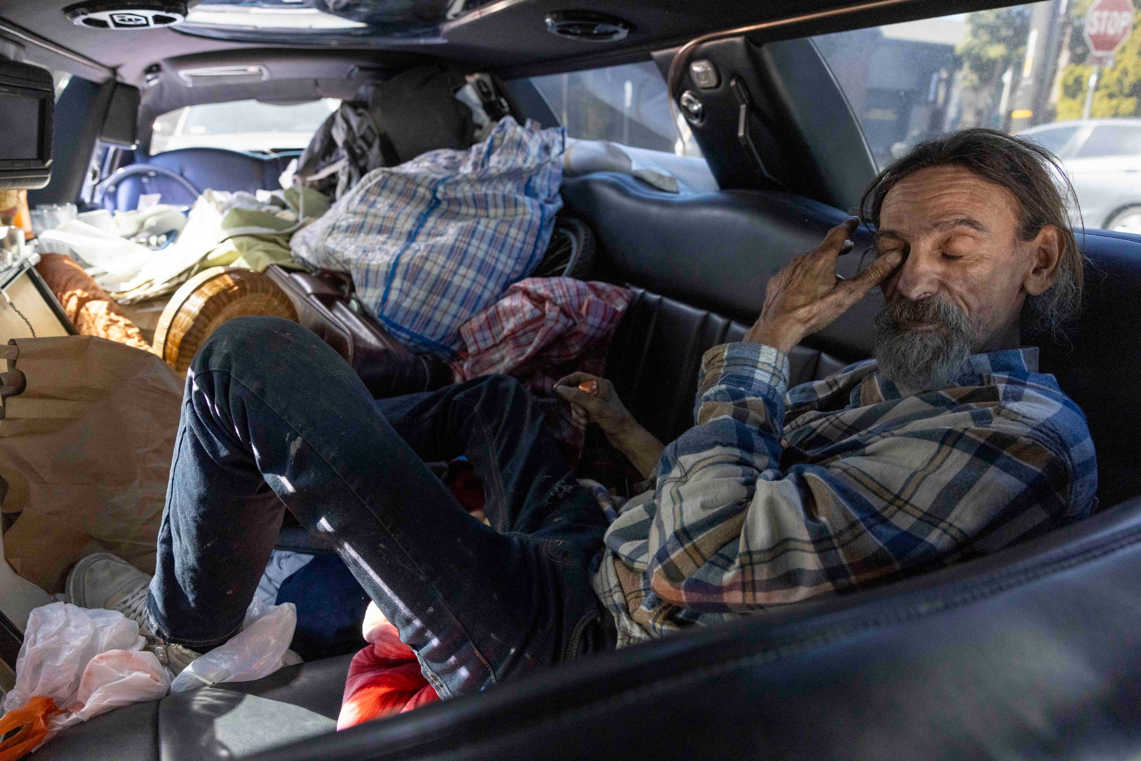 A man with a beard, wearing a plaid shirt, rests inside a cluttered car, surrounded by bags and personal belongings, appearing to be in a thoughtful state.