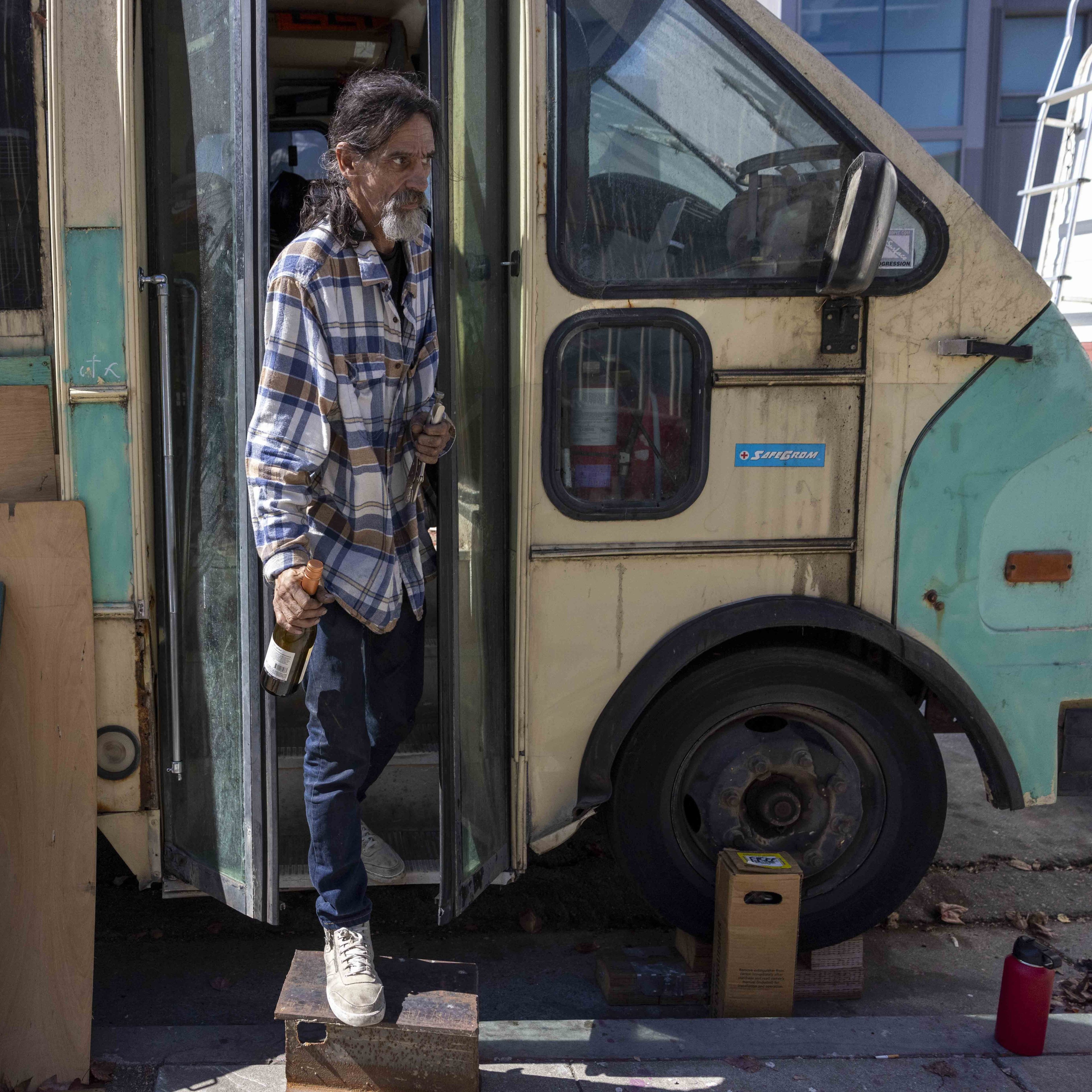 A man in a plaid shirt steps out of an old, worn bus. He holds a bottle in one hand, using a block as a step on a street with cardboard boxes nearby.