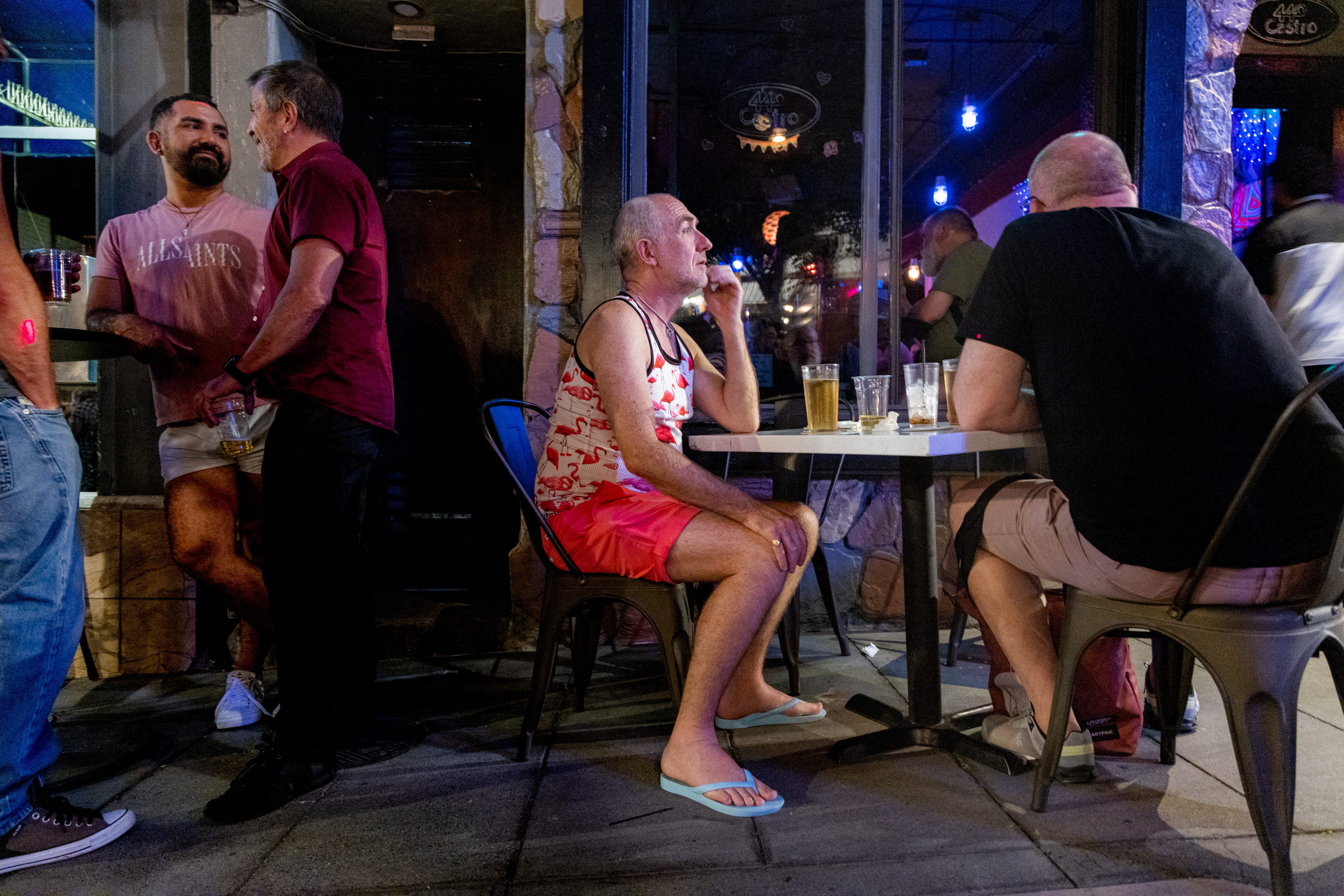 Two men stand chatting by a wall, while another man sits at a table outside a bar, wearing a flamingo tank top, shorts, and flip-flops.