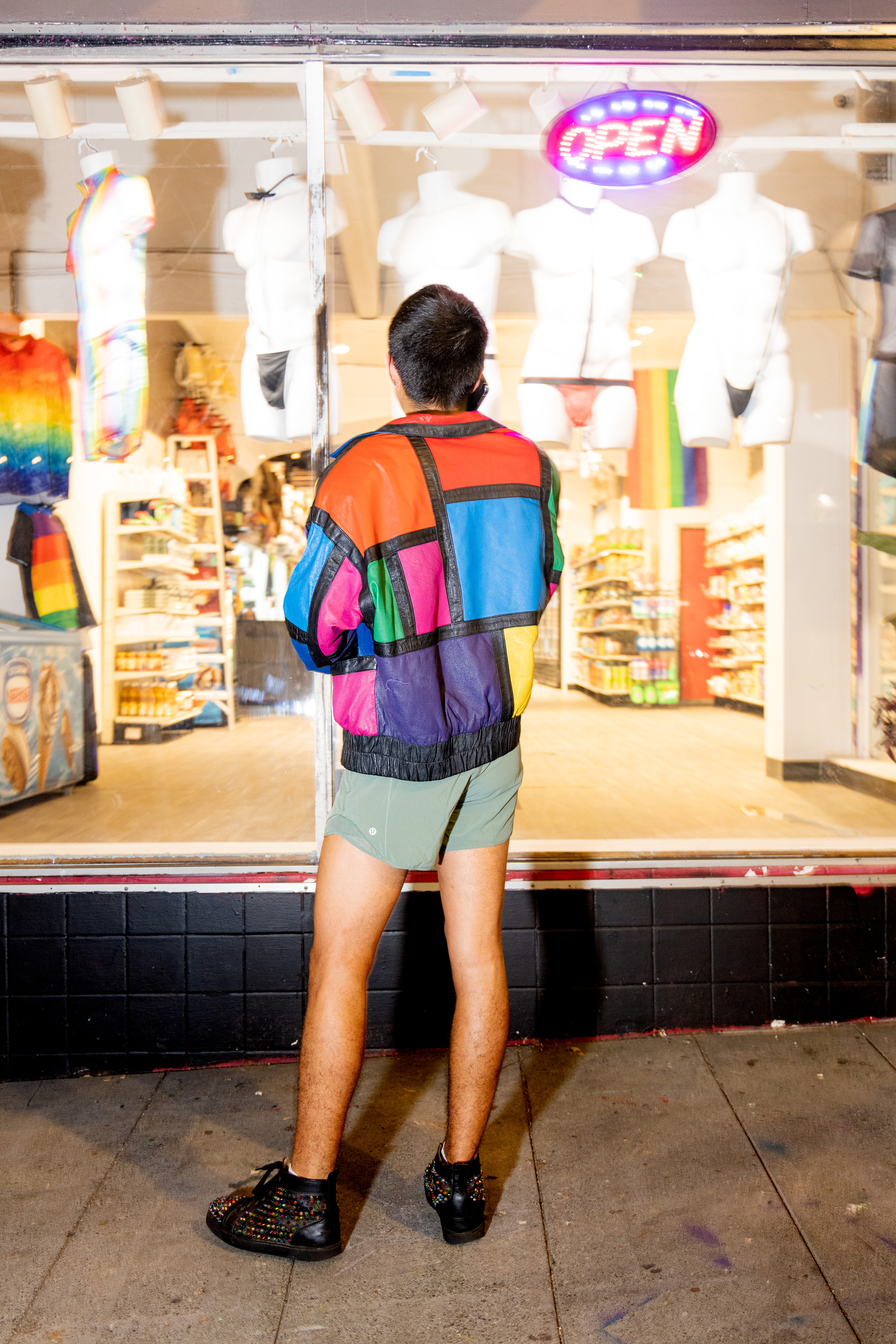 A person in a colorful, patchwork jacket and shorts stands in front of a shop window displaying mannequins in swimwear. A glowing &quot;Open&quot; sign is visible above.