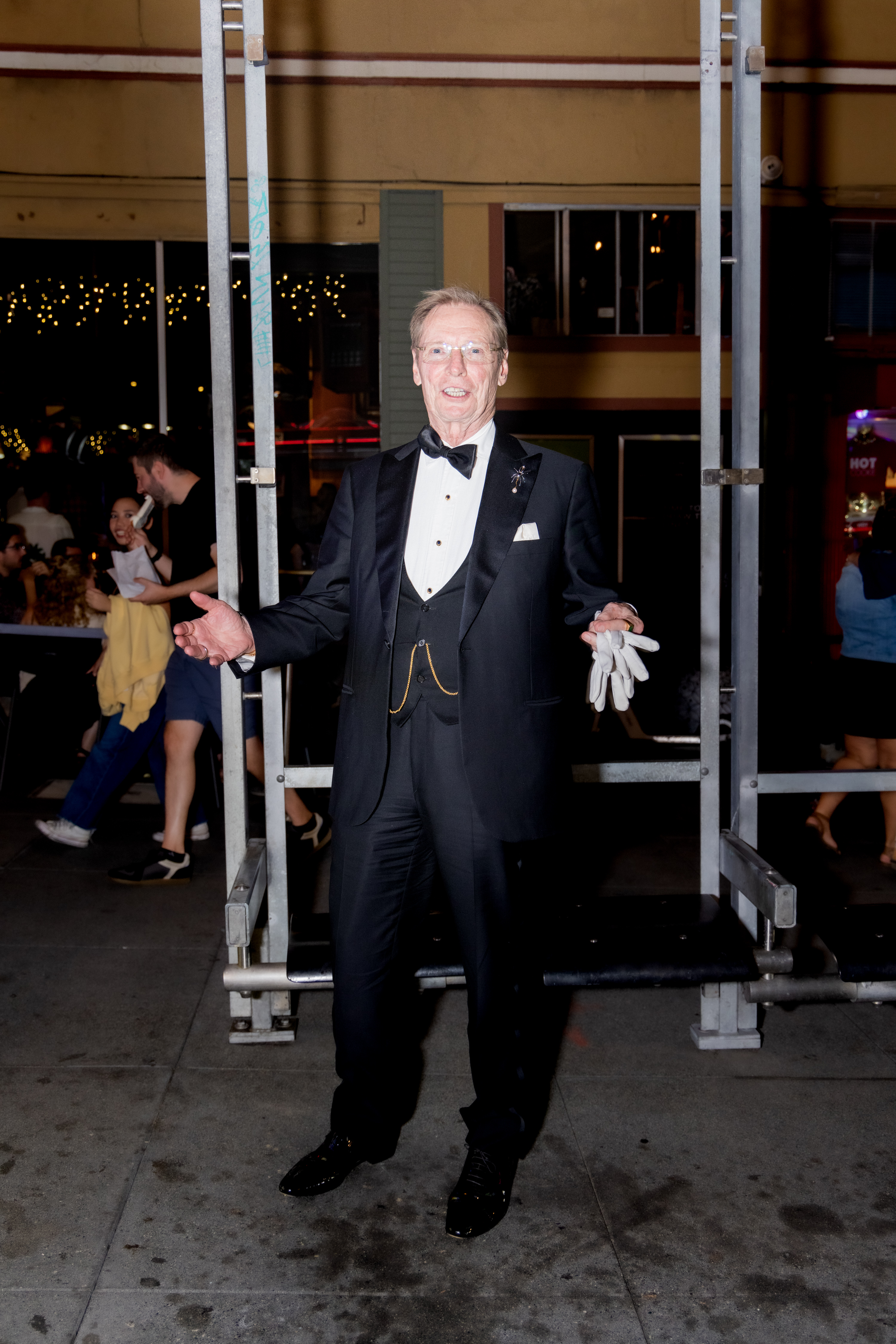 A man in a tuxedo stands smiling on a street at night, holding white gloves. Behind him, people are socializing under string lights.