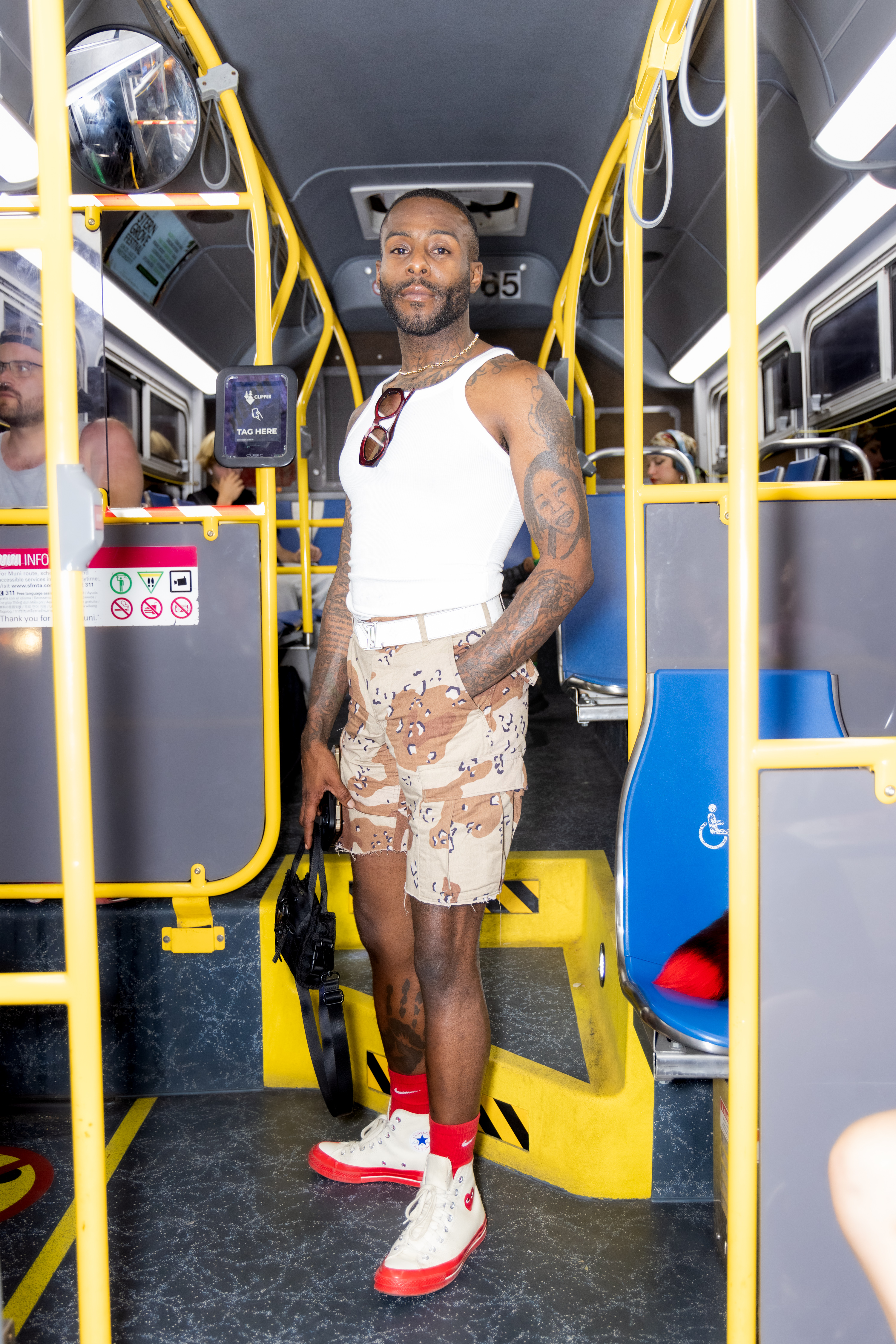 A person on a bus wears a white tank top, patterned shorts, and red socks with high-top sneakers. They have tattoos and hold a black bag, standing confidently.