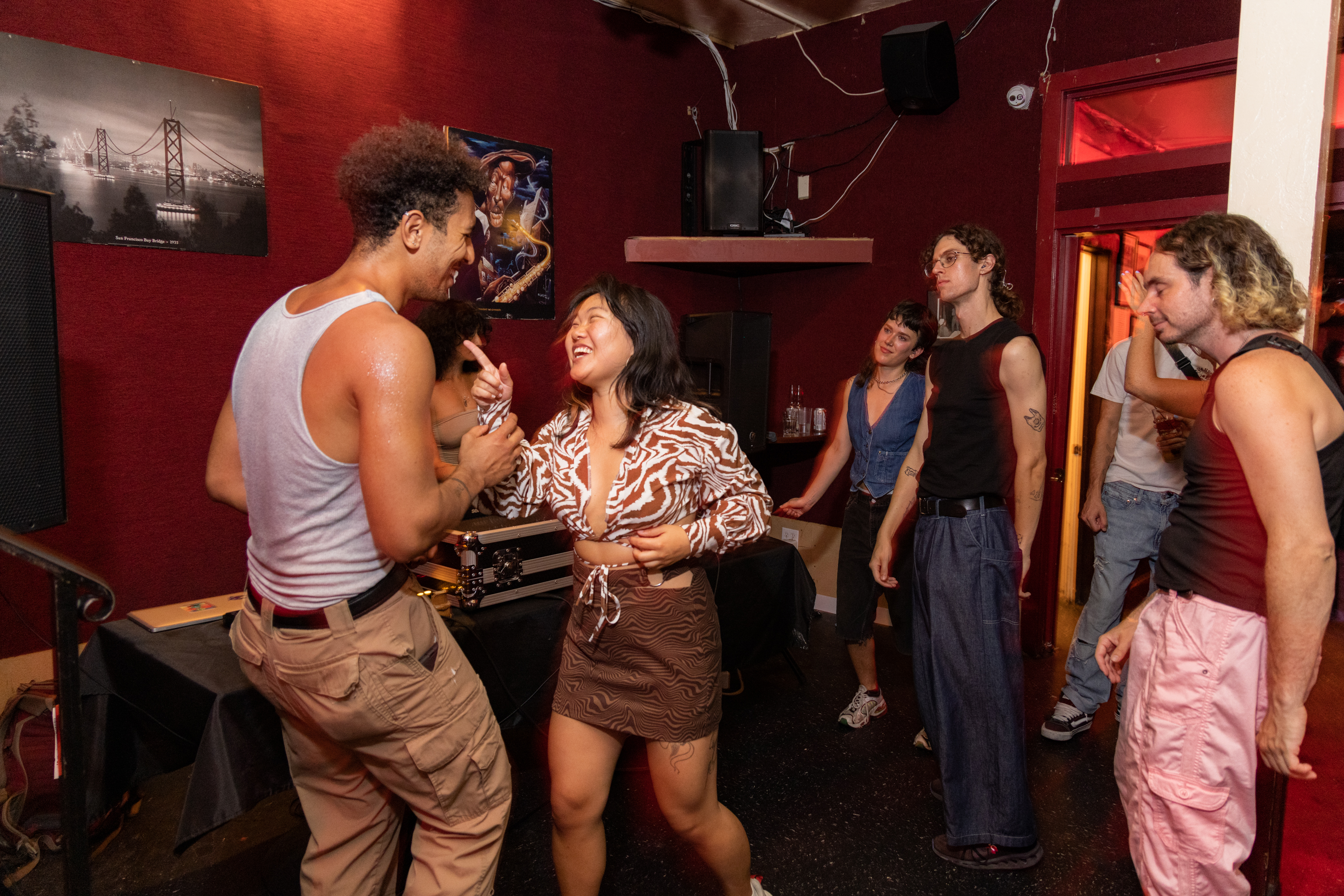 A lively group of people are socializing indoors. Two individuals in the center are holding hands and smiling. The room has dark red walls and artwork on the wall.