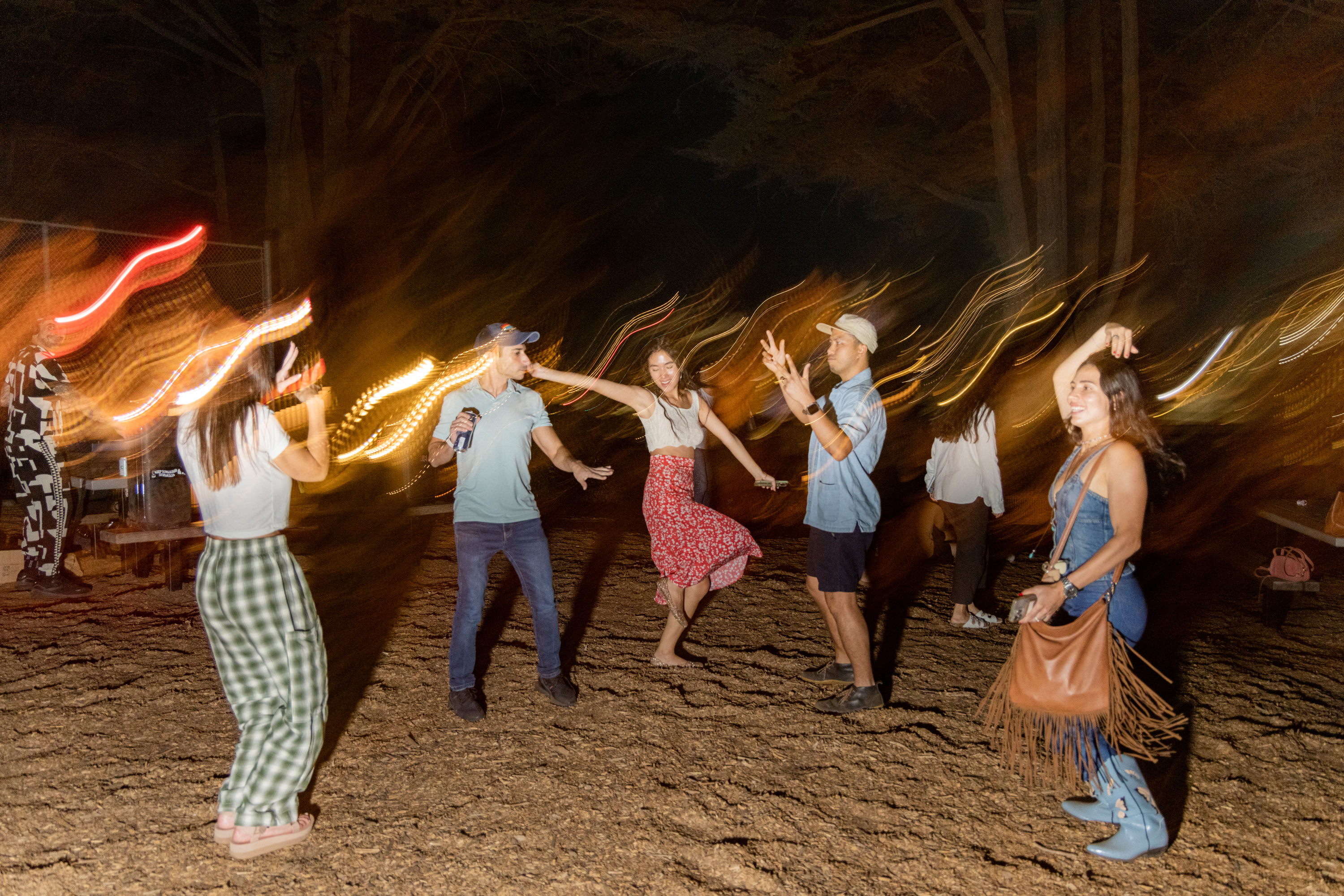 A group of people are dancing outside at night, with blurred motion and light trails around them. The setting is casual, with everyone in relaxed attire.