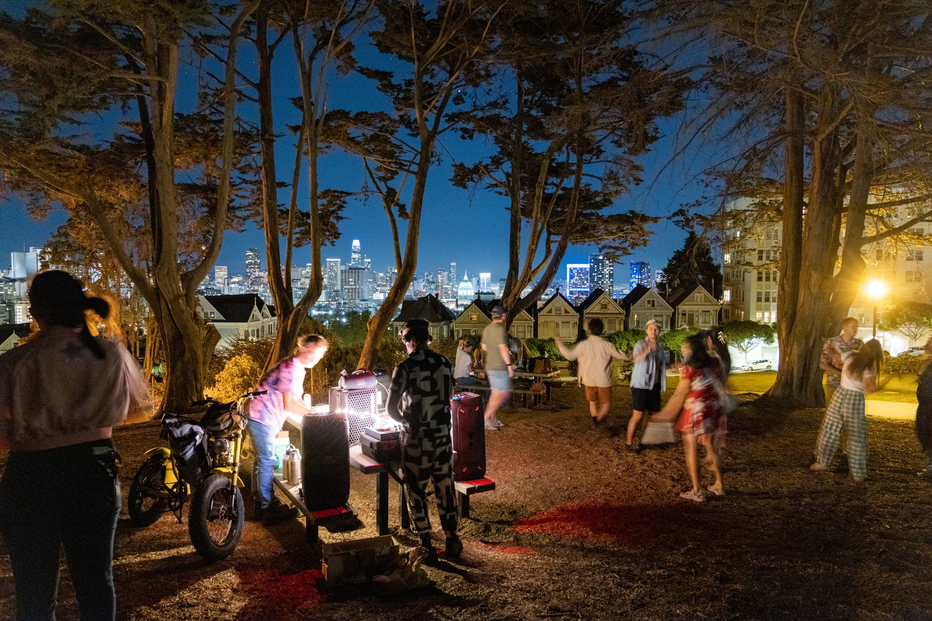 People gather in a park at night with lit tables and a city skyline in the background. Trees frame the scene, and there's a mix of standing and walking individuals.