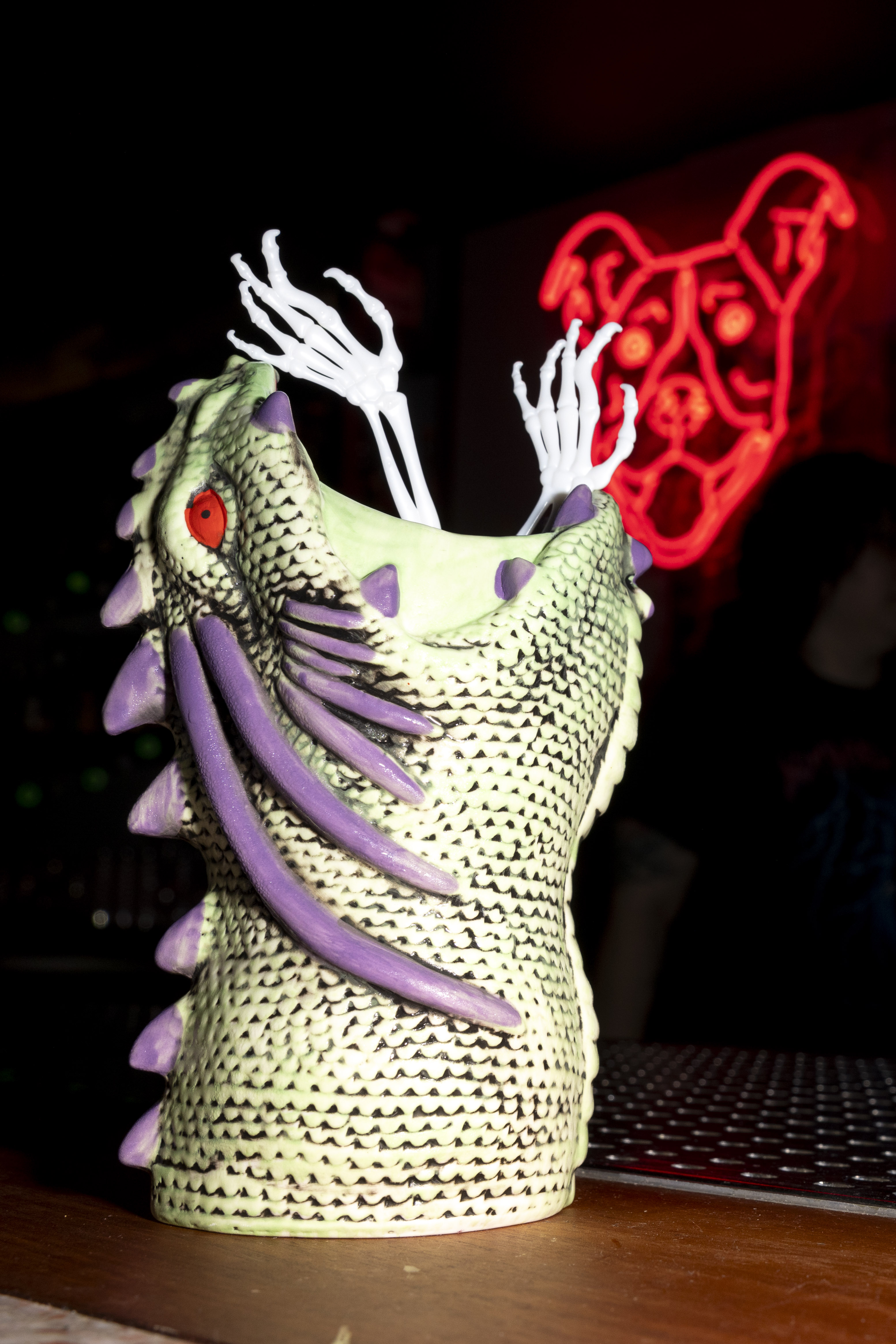 A textured, dragon-like ceramic cup with purple accents and white skeletal hands protruding. A neon red dog face sign glows in the background.