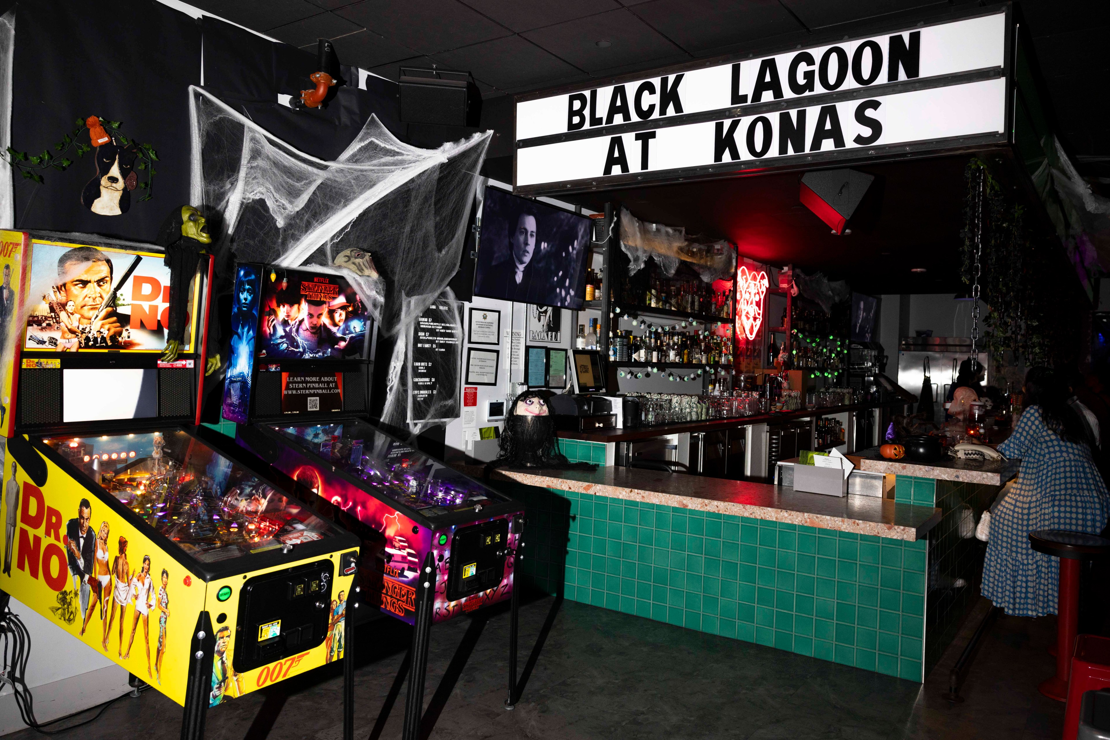 The image shows a bar with green tiles, two pinball machines, spooky decorations, and a sign saying &quot;BLACK LAGOON AT KONAS.&quot; There's a person at the bar.