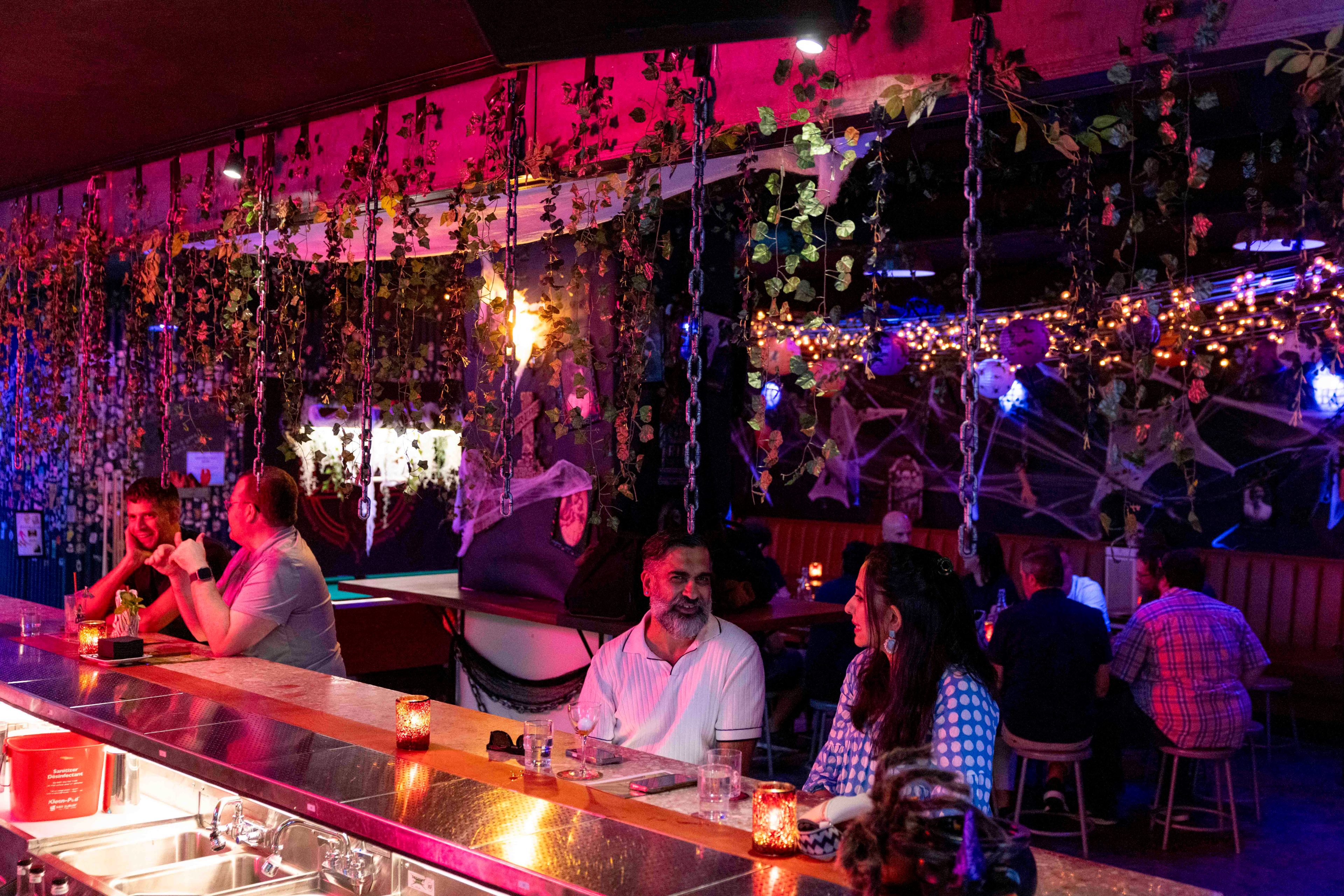 A vibrant bar with colorful lighting, hanging plants, and chains. People are seated along the bar and booths, chatting amidst dim, festive decor.