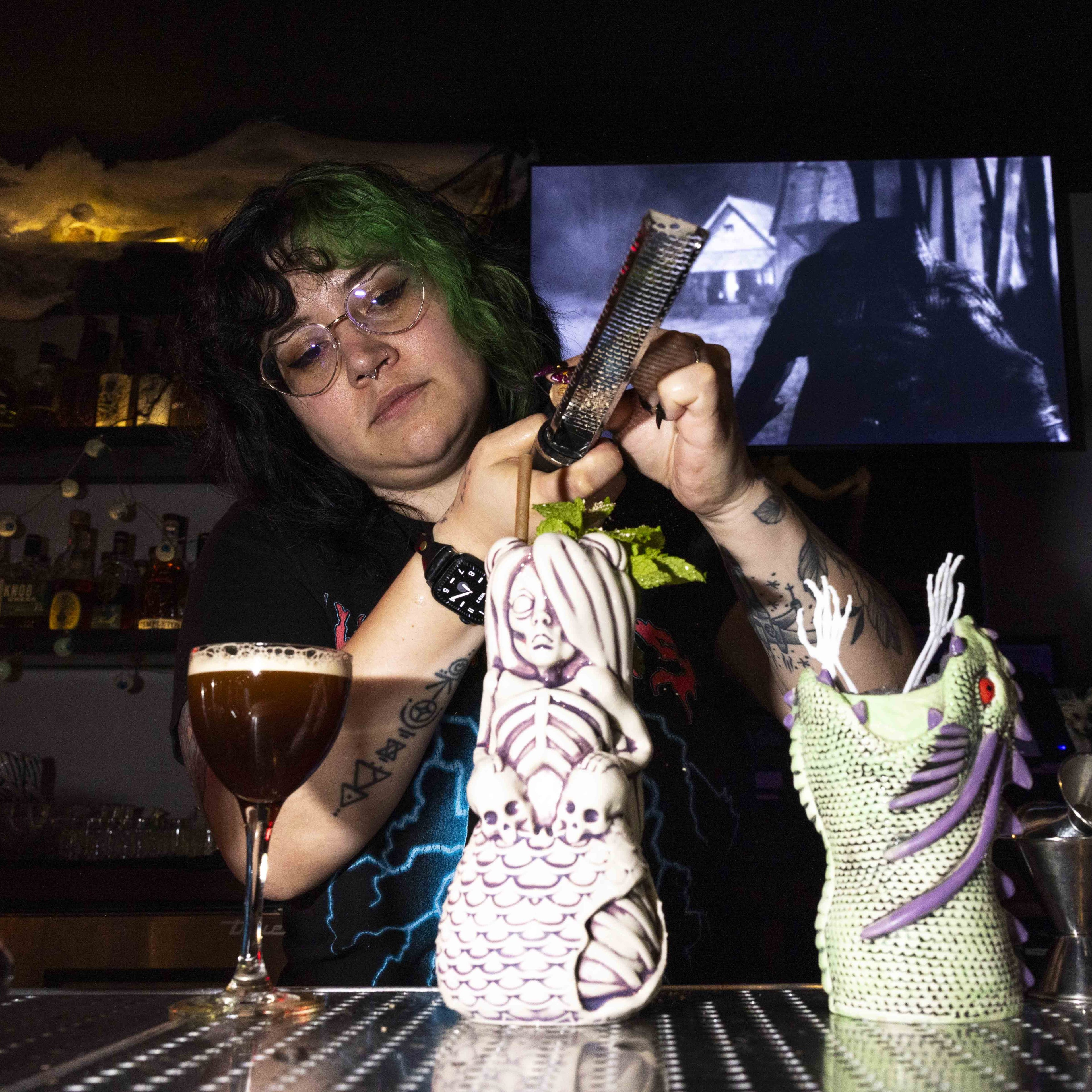 A person with green and black hair and tattoos is grating something over a decorative drink in a mermaid-themed cup. A cocktail and dragon cup are nearby.