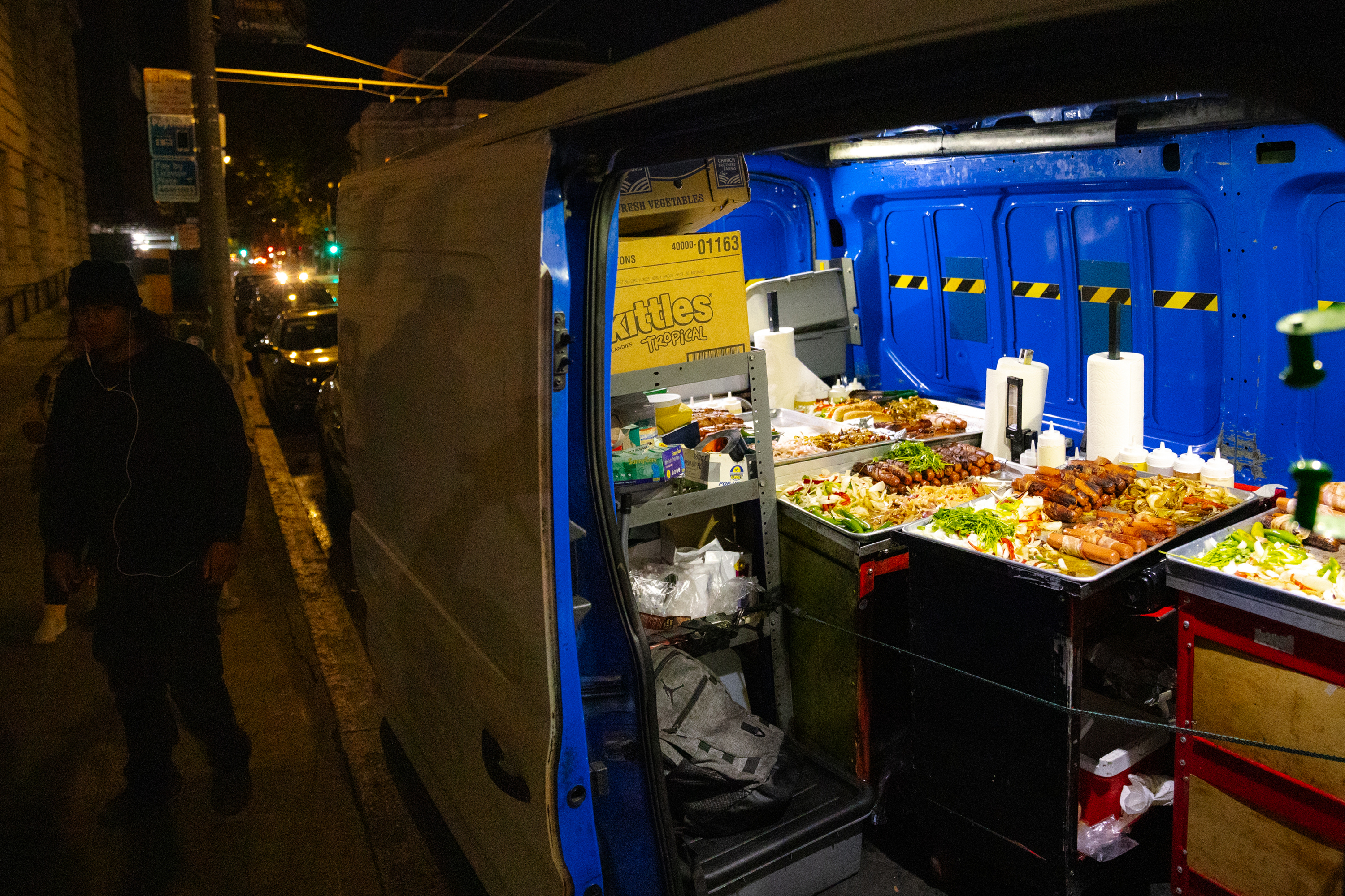 A nighttime street scene shows a van with its side door open, revealing colorful trays of food inside. A person walks by on the sidewalk nearby.