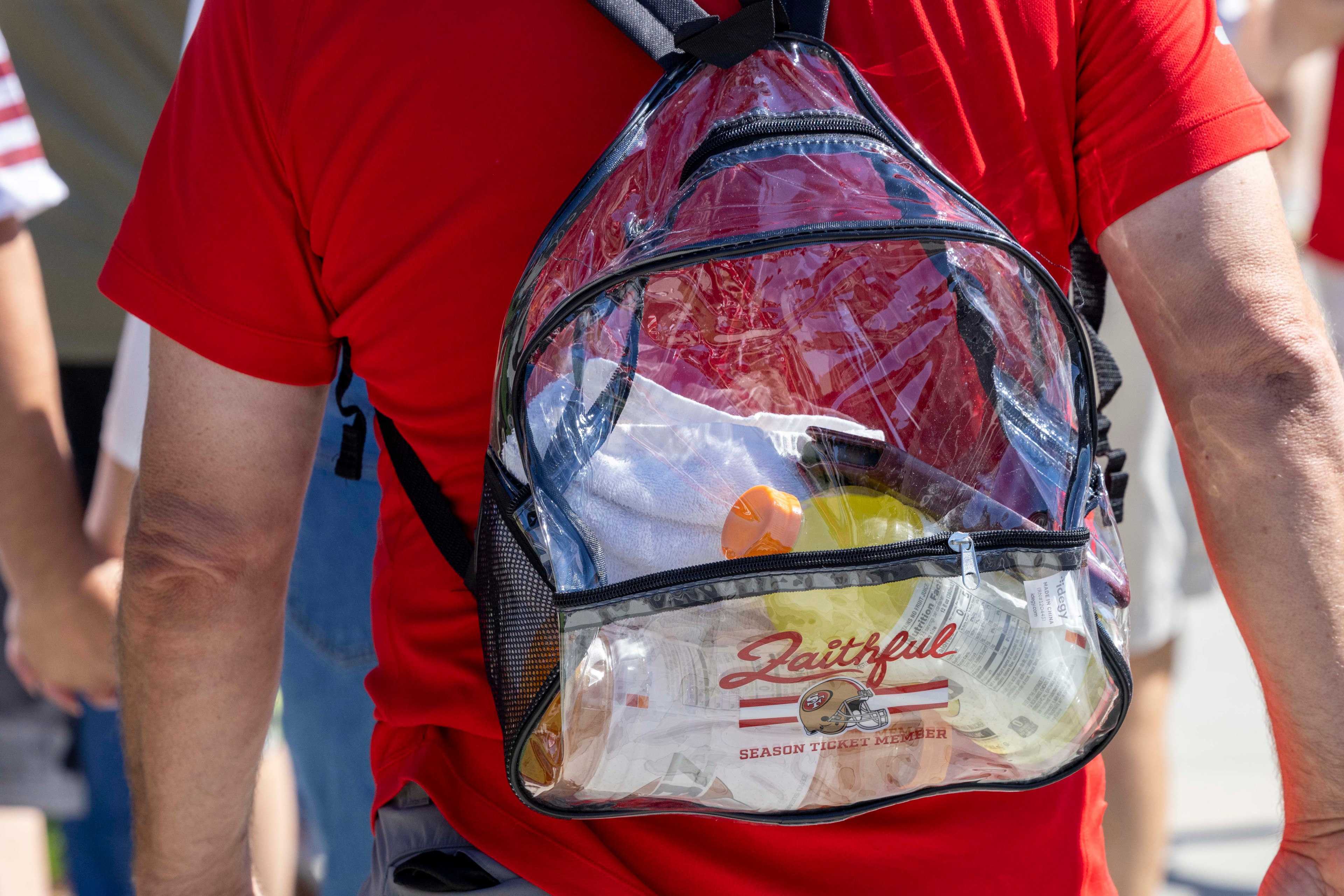 A person in a red shirt is wearing a clear backpack, revealing a water bottle, towel, and items labeled &quot;Faithful&quot; with a sports team logo.