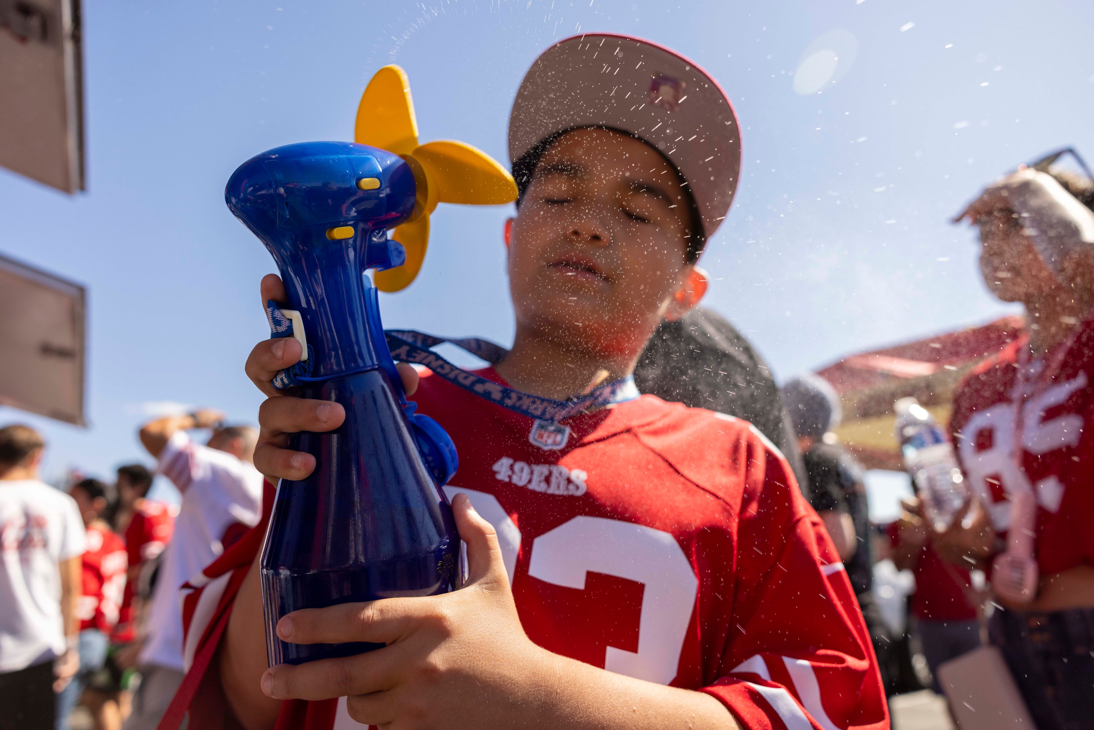 A child wearing a red sports jersey and cap holds a blue misting fan, spraying themselves with water. There's a sunny crowd in the background, also dressed in red.