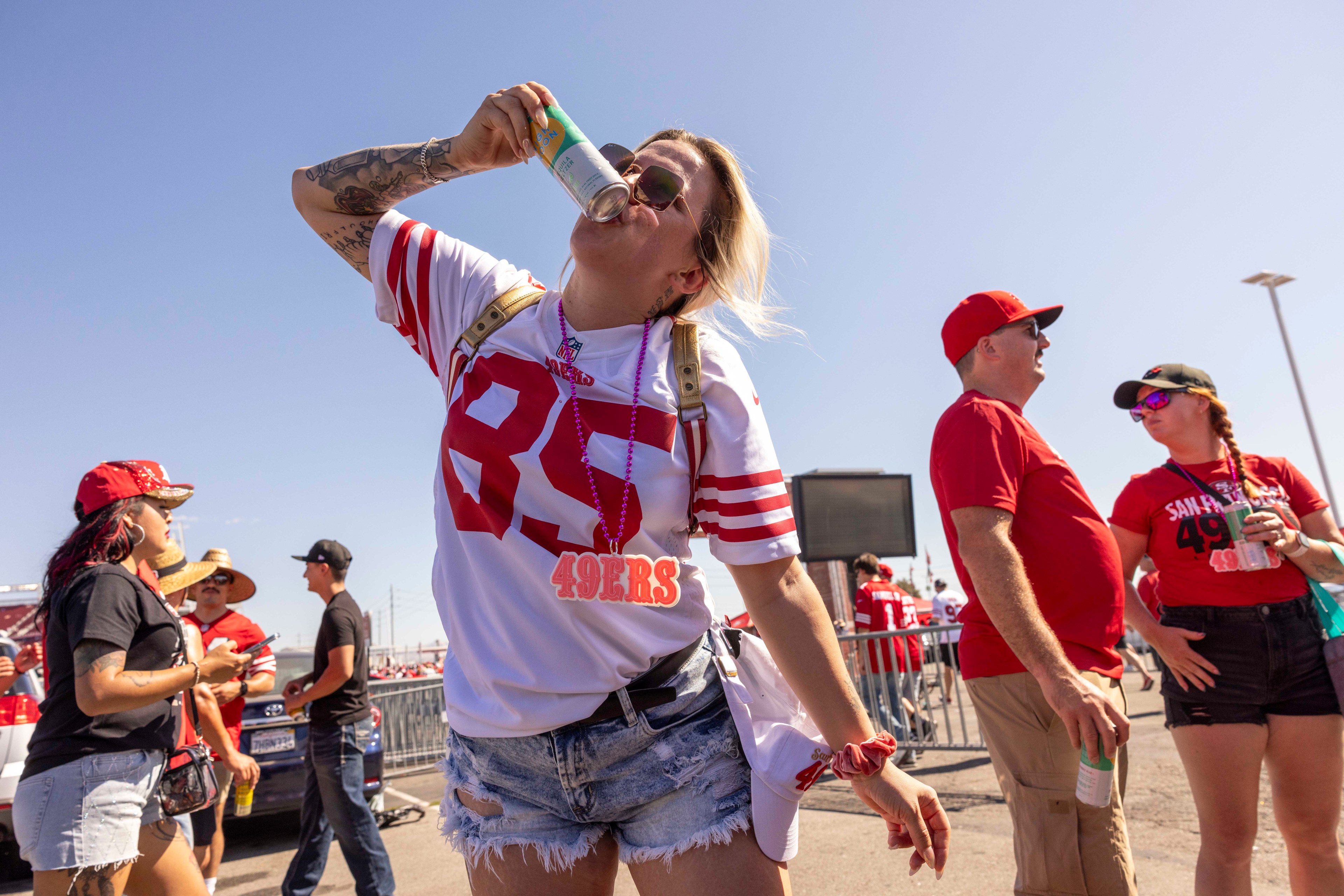 A woman wearing a 49ers jersey and jean shorts drinks from a can at an outdoor event, surrounded by people in red clothing, under a clear blue sky.