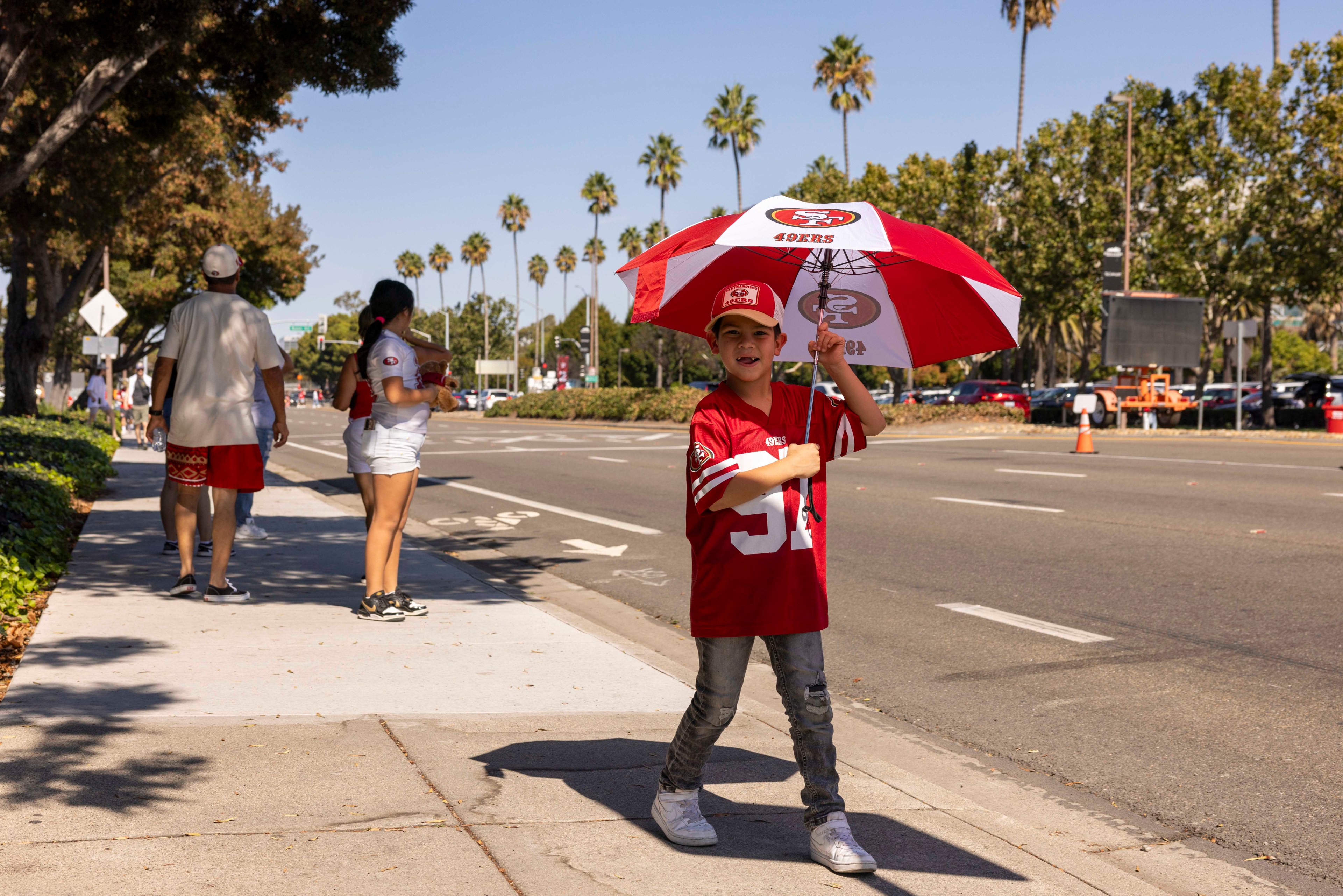 A child in a red jersey and cap holds a matching umbrella on a sunny sidewalk, with palm trees and parked cars in the background. Nearby, people wear similar sports attire.