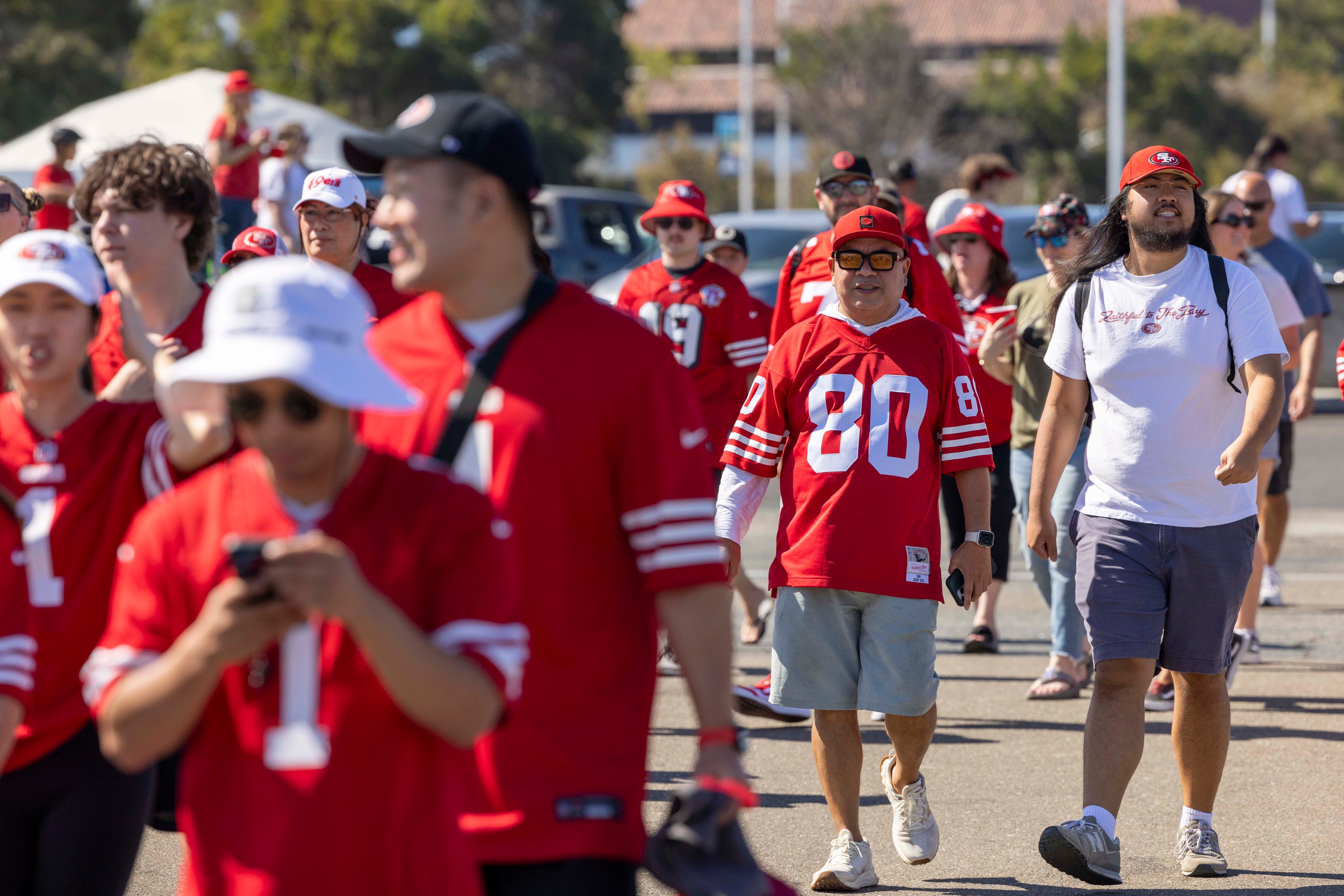 A group of people in red sports jerseys and hats walk outdoors on a sunny day. Some are checking their phones, while others chat and smile.