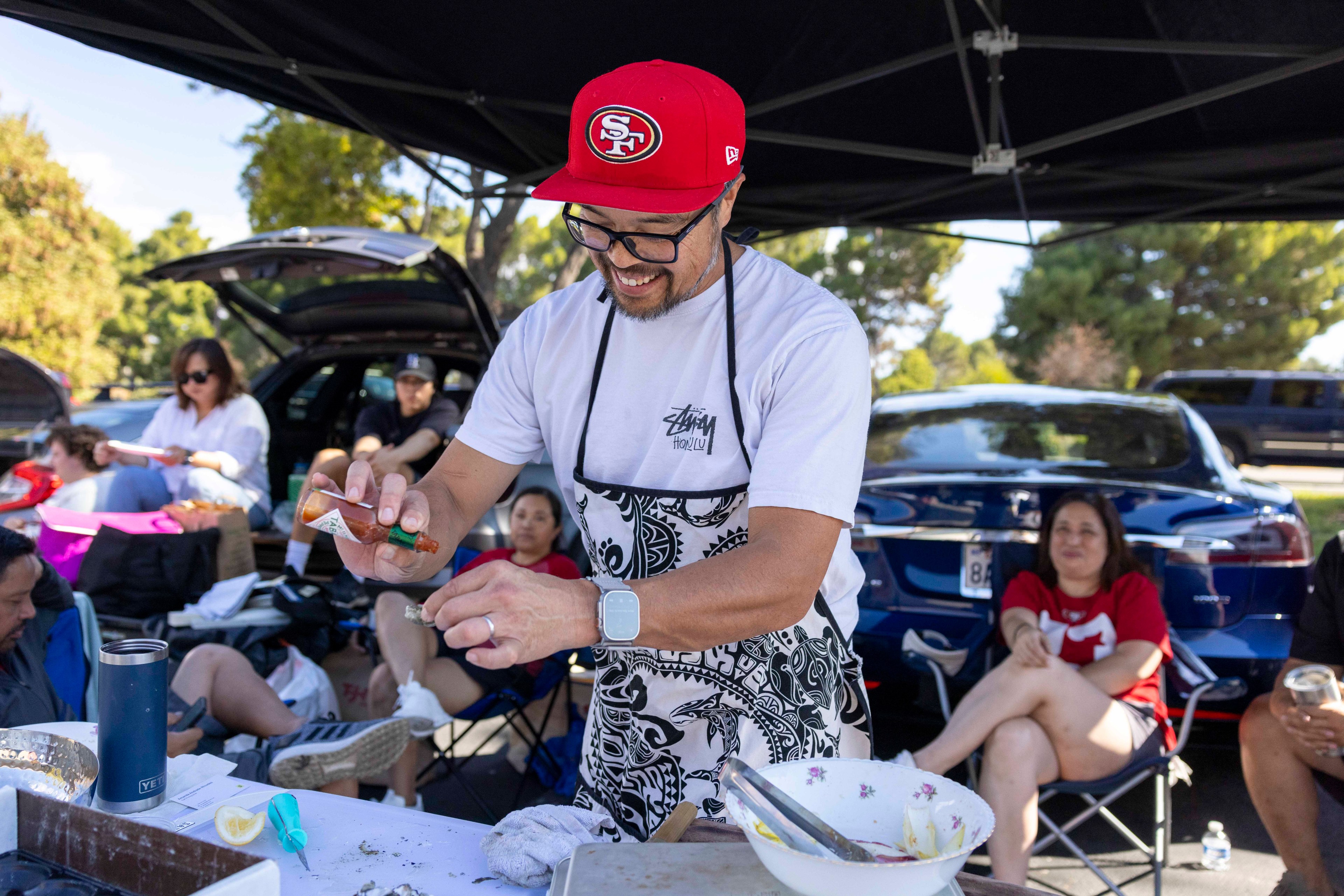 A man in a red cap and patterned apron prepares food, smiling under a canopy. Behind him, people relax in folding chairs near parked cars.