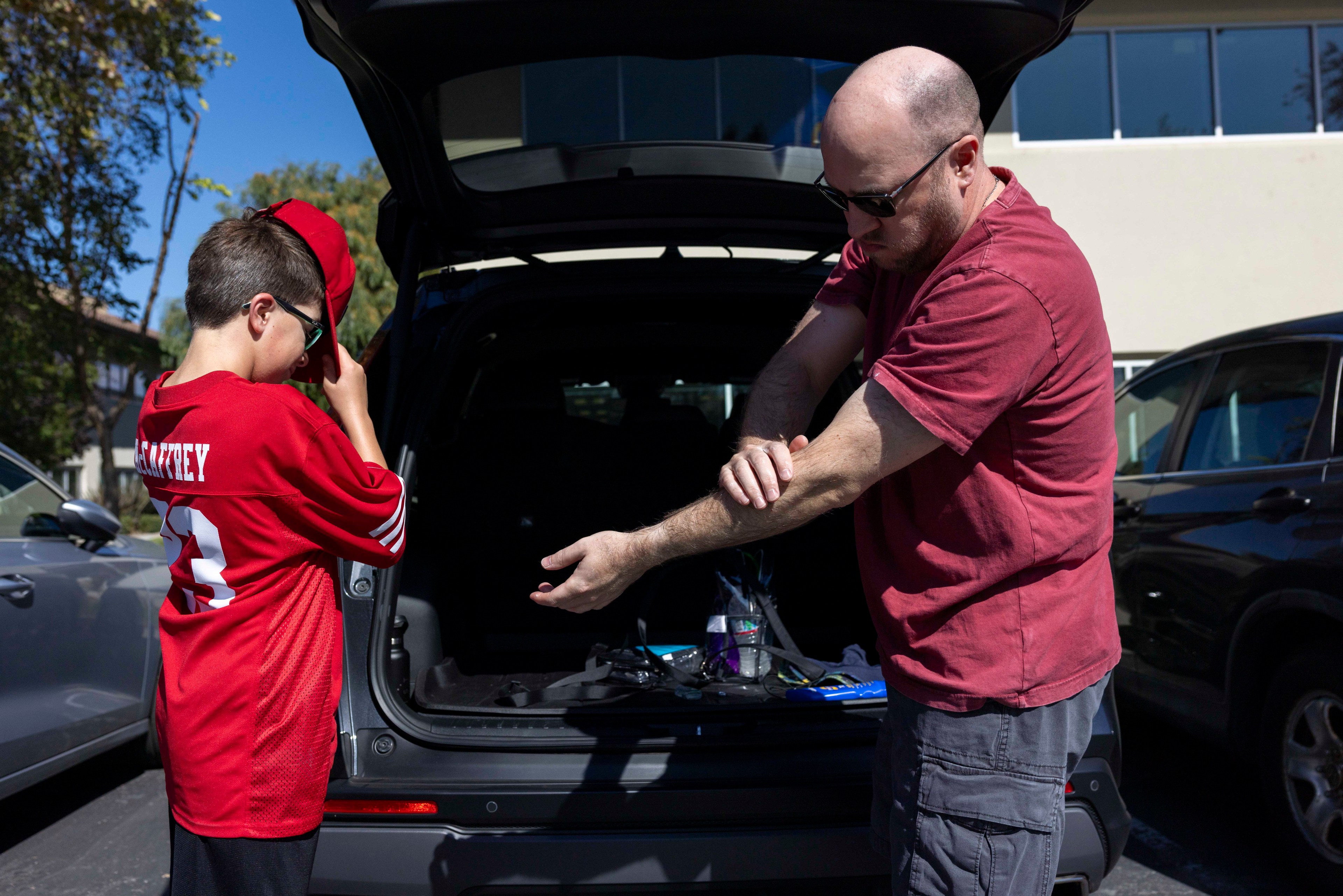 A man in sunglasses applies sunscreen on his arm, standing by an SUV with its trunk open. A boy in a red sports jersey adjusts his cap nearby.