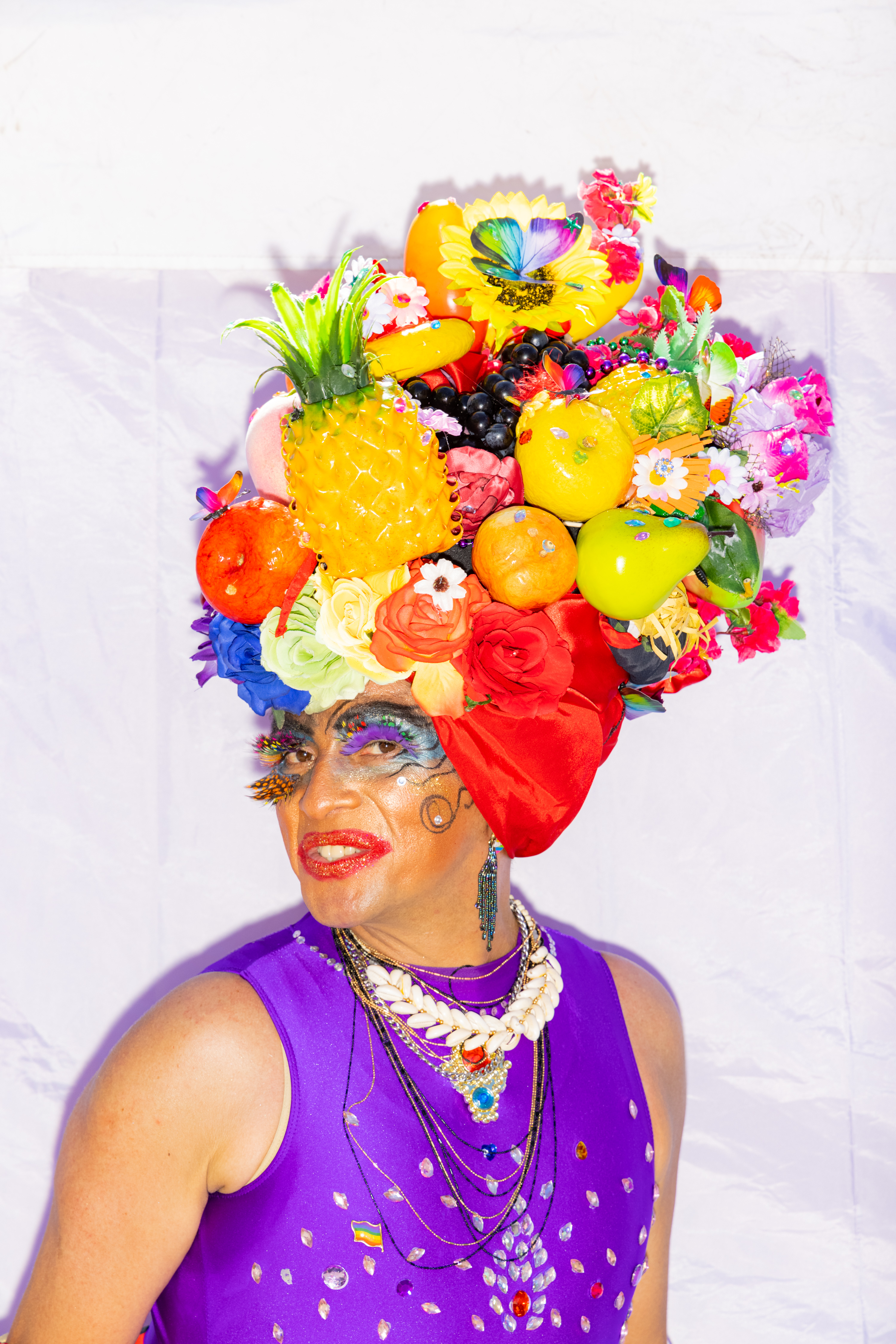 A person wears a vibrant fruit and flower headdress, colorful makeup, and a purple top adorned with sparkles and layered necklaces.