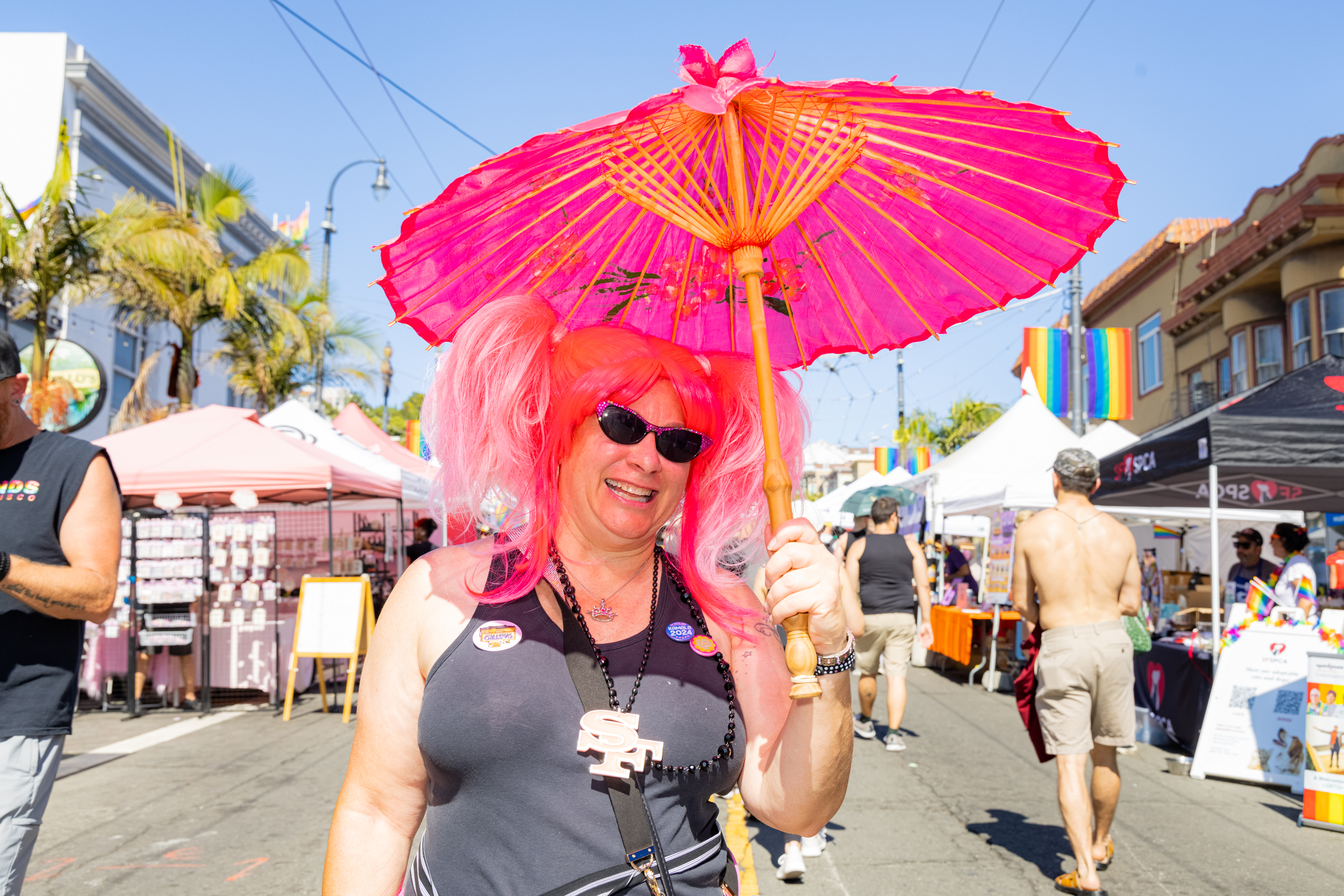 A person in a vibrant pink wig and sunglasses holds a bright pink parasol. They are smiling at an outdoor event with tents and people in the background.